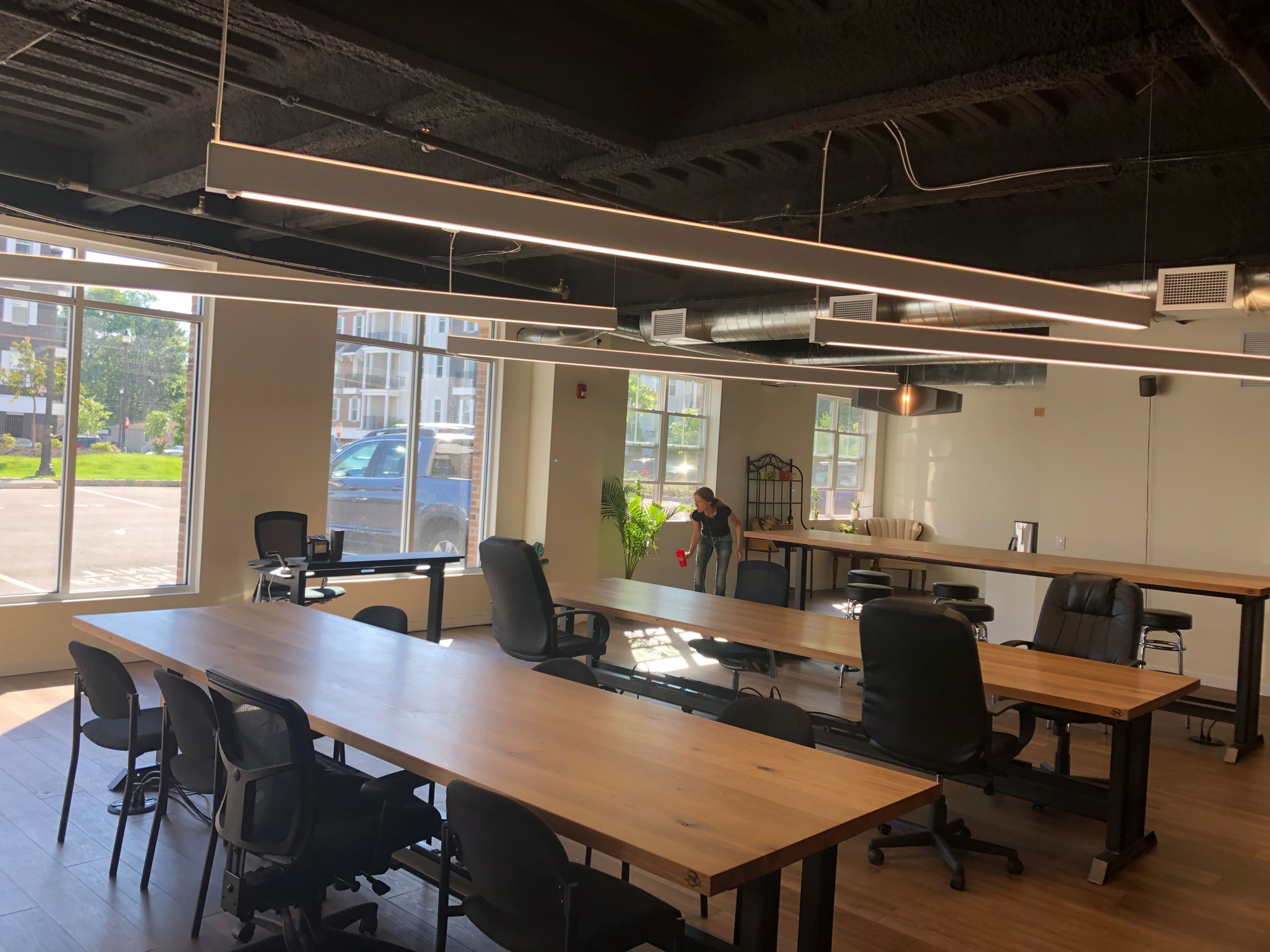 A modern office space features long wooden tables, black ergonomic chairs, and large windows allowing natural light to enter.
