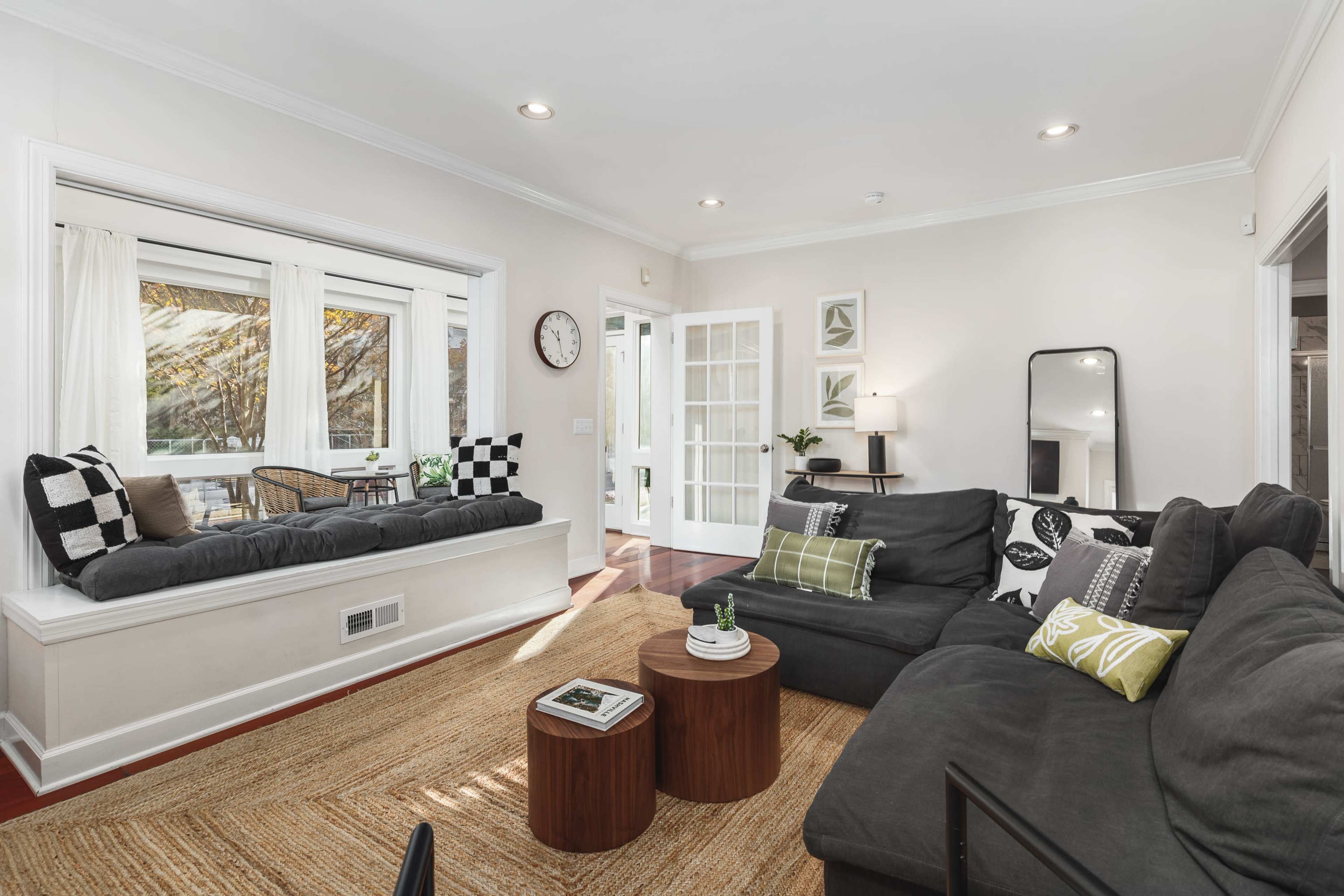 A cozy living room features a sectional sofa, a bay window with seating, two wooden side tables, and a mix of decorative pillows.