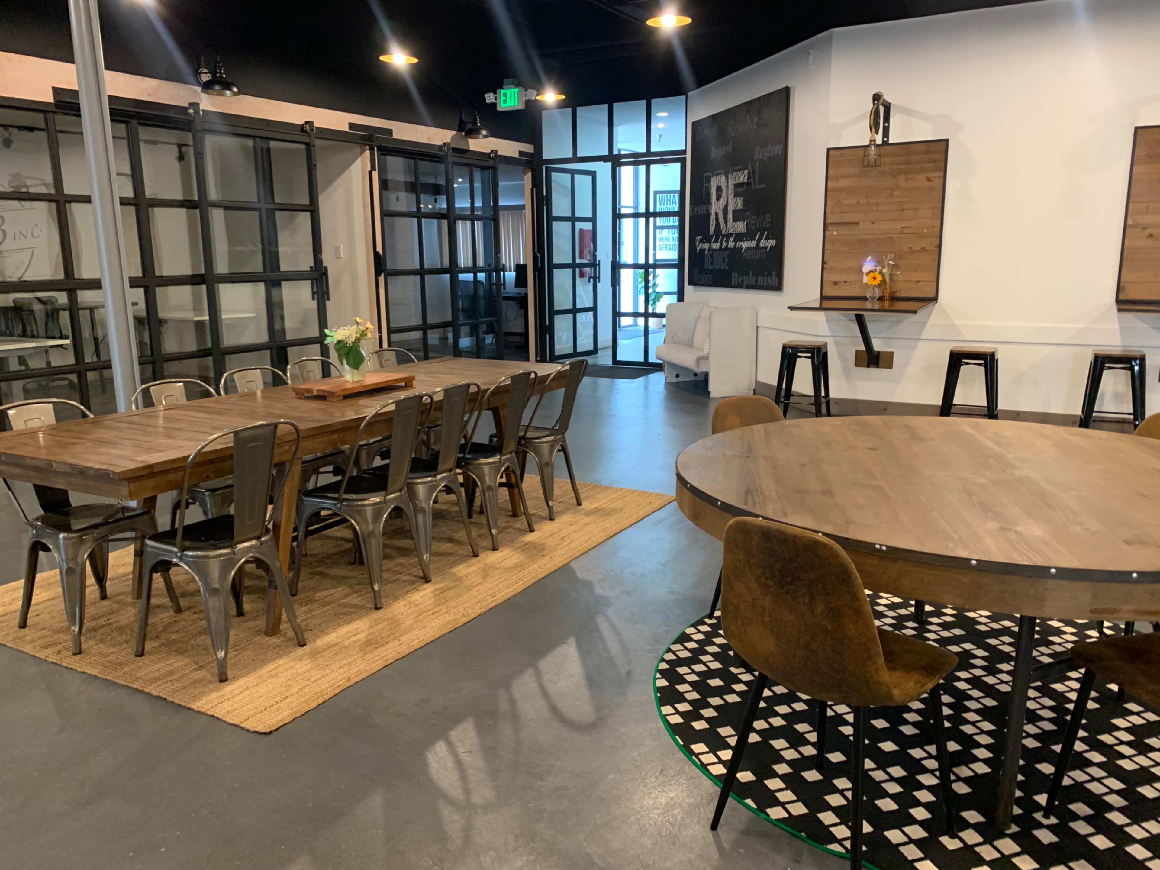 The image shows a modern interior space featuring a long wooden dining table with metal chairs, a round wooden table with upholstered chairs, and large glass doors opening to an exterior area.