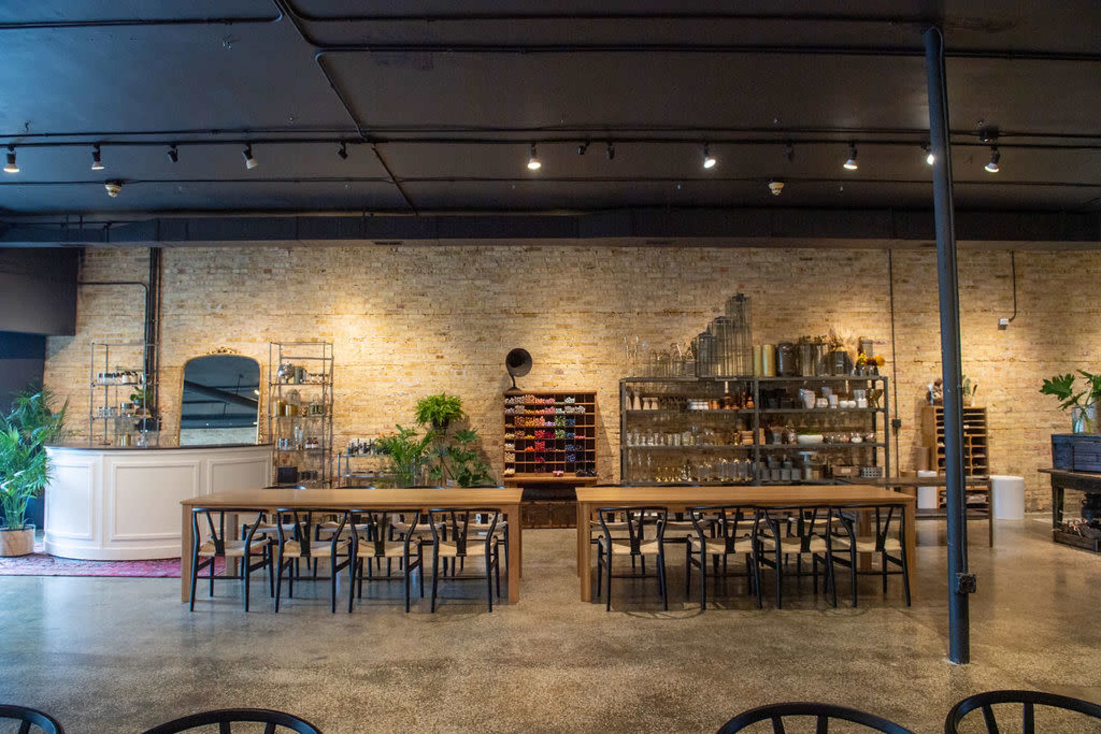 A spacious interior of a restaurant featuring a wooden dining table, metal chairs, and a brick wall with shelves displaying various glassware and plants.
