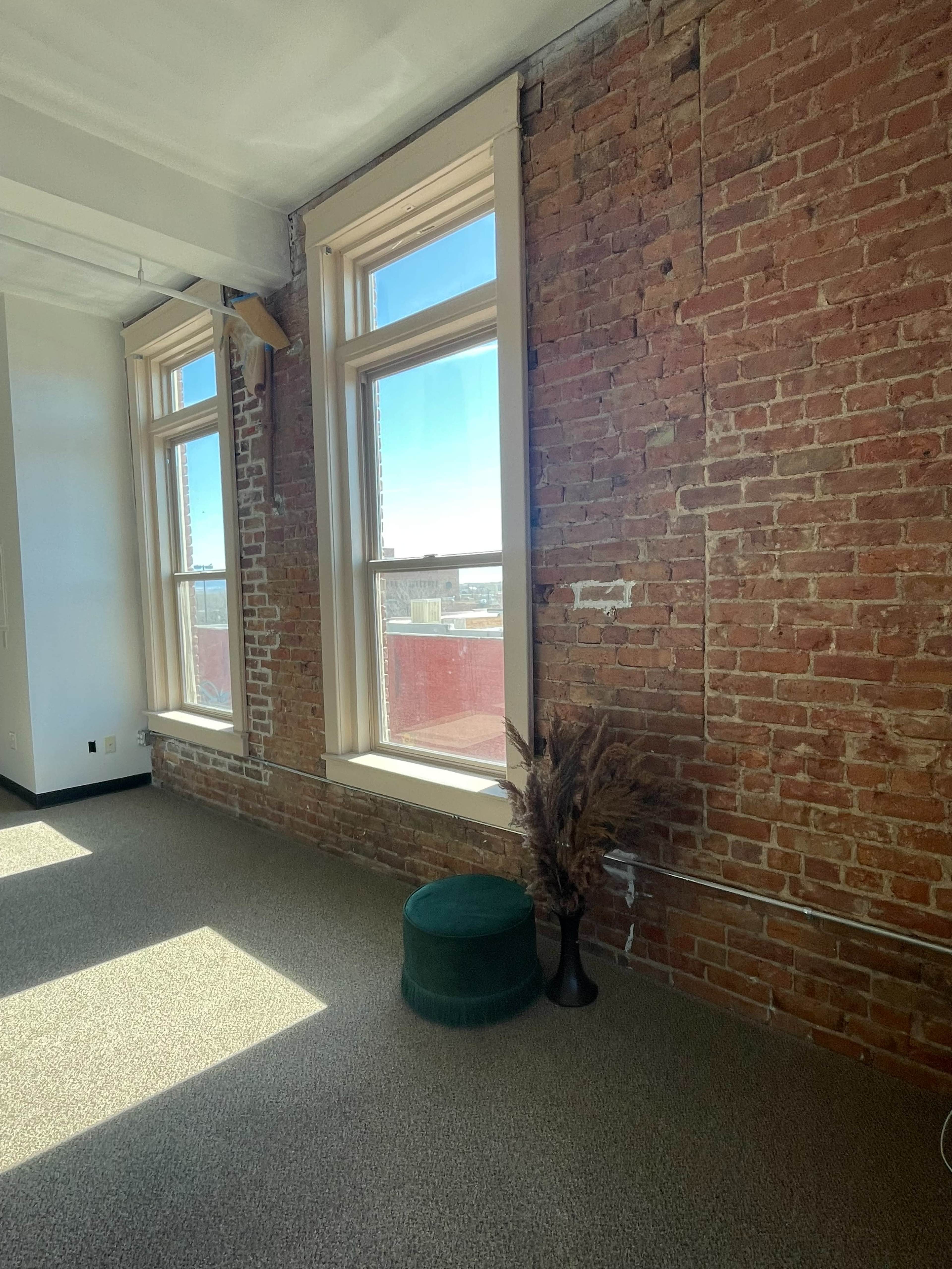 The image shows a bright room with two large windows, exposed brick walls, and a small green stool next to a decorative plant.