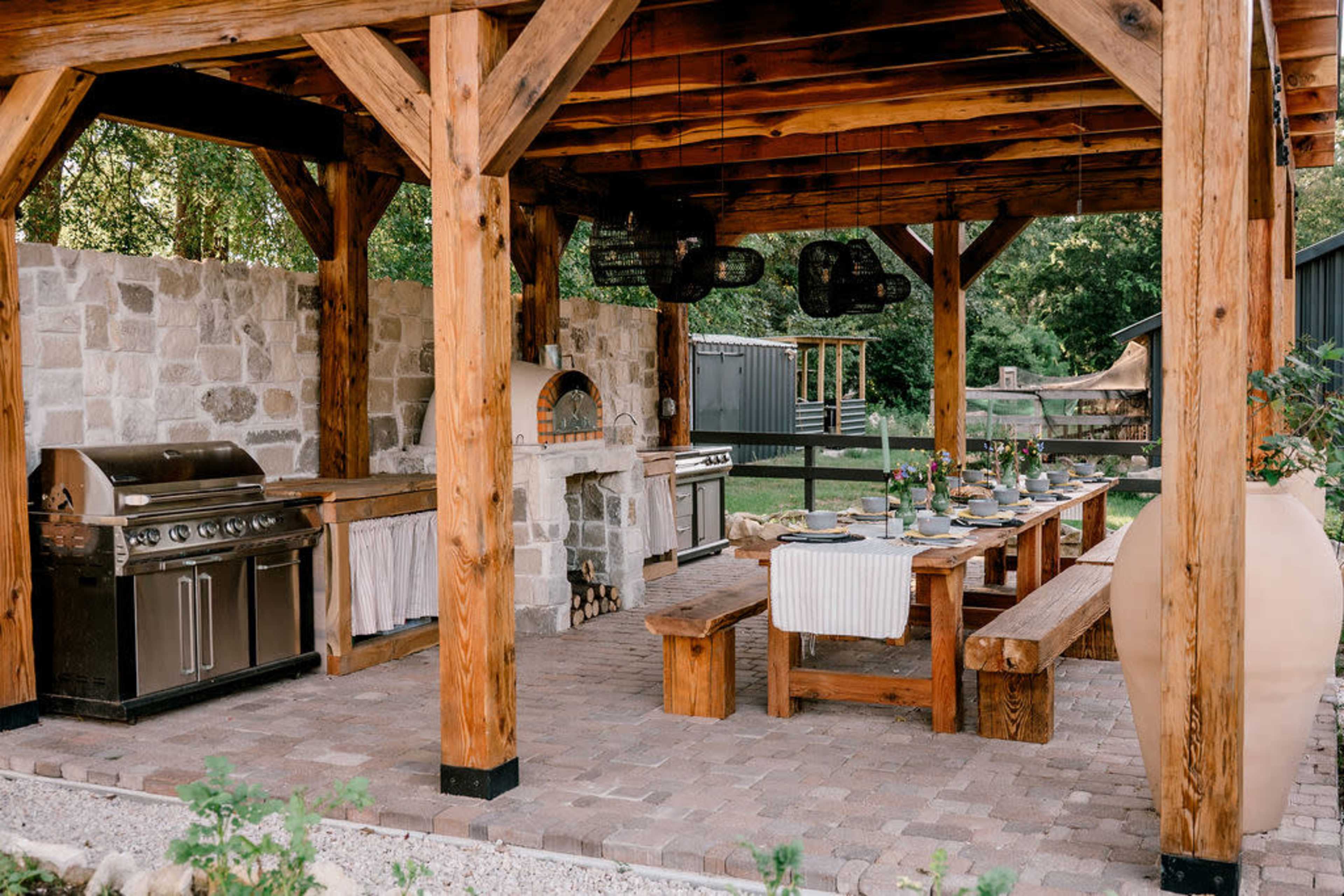 The image shows a wooden pergola with a stone wall, featuring an outdoor kitchen setup that includes a grill, an oven, and a long dining table with benches arranged underneath.