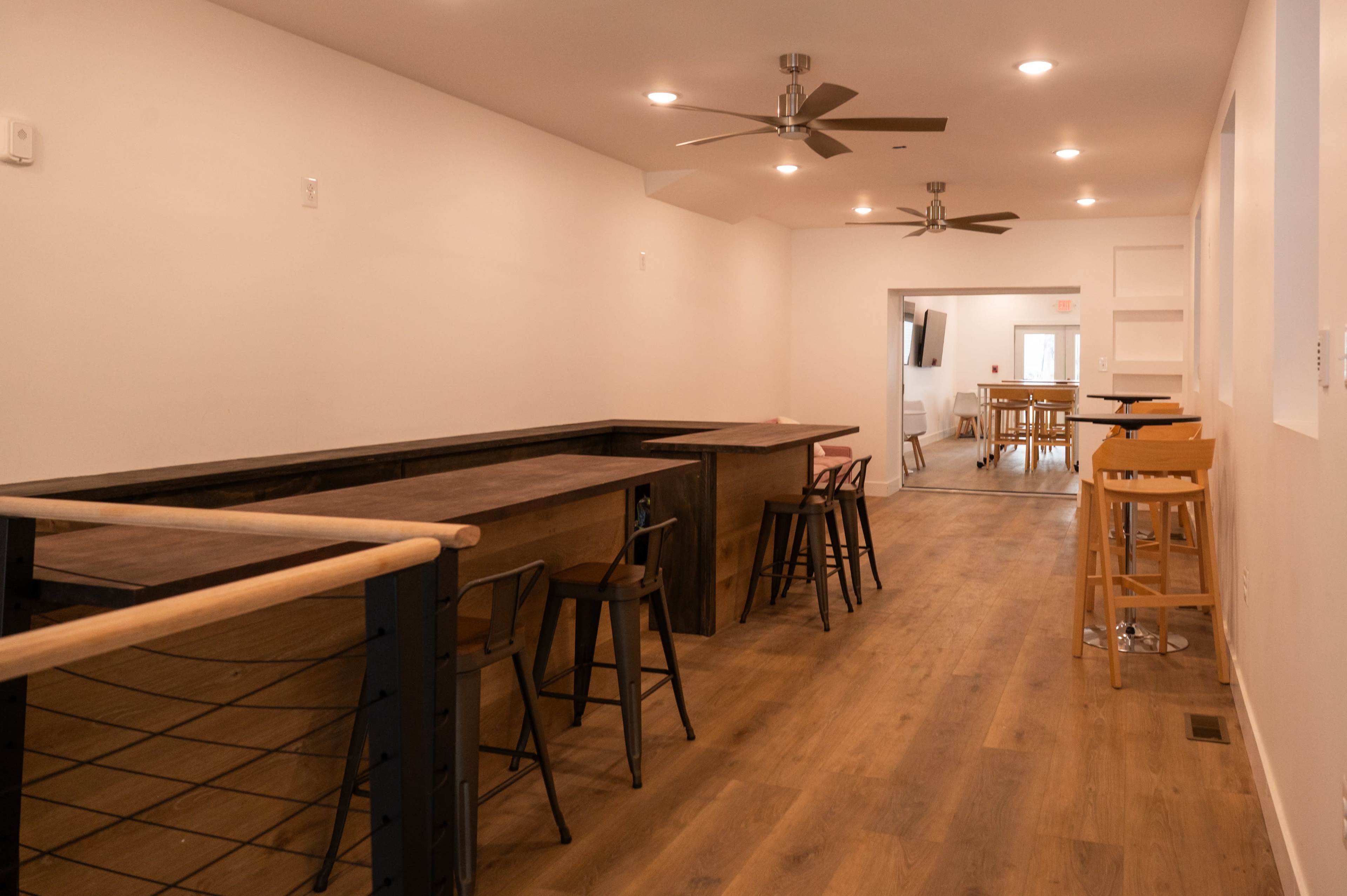 The image shows a modern, open space featuring wooden countertops, a mixture of seating options, and ceiling fans, with natural light coming from windows at the far end.
