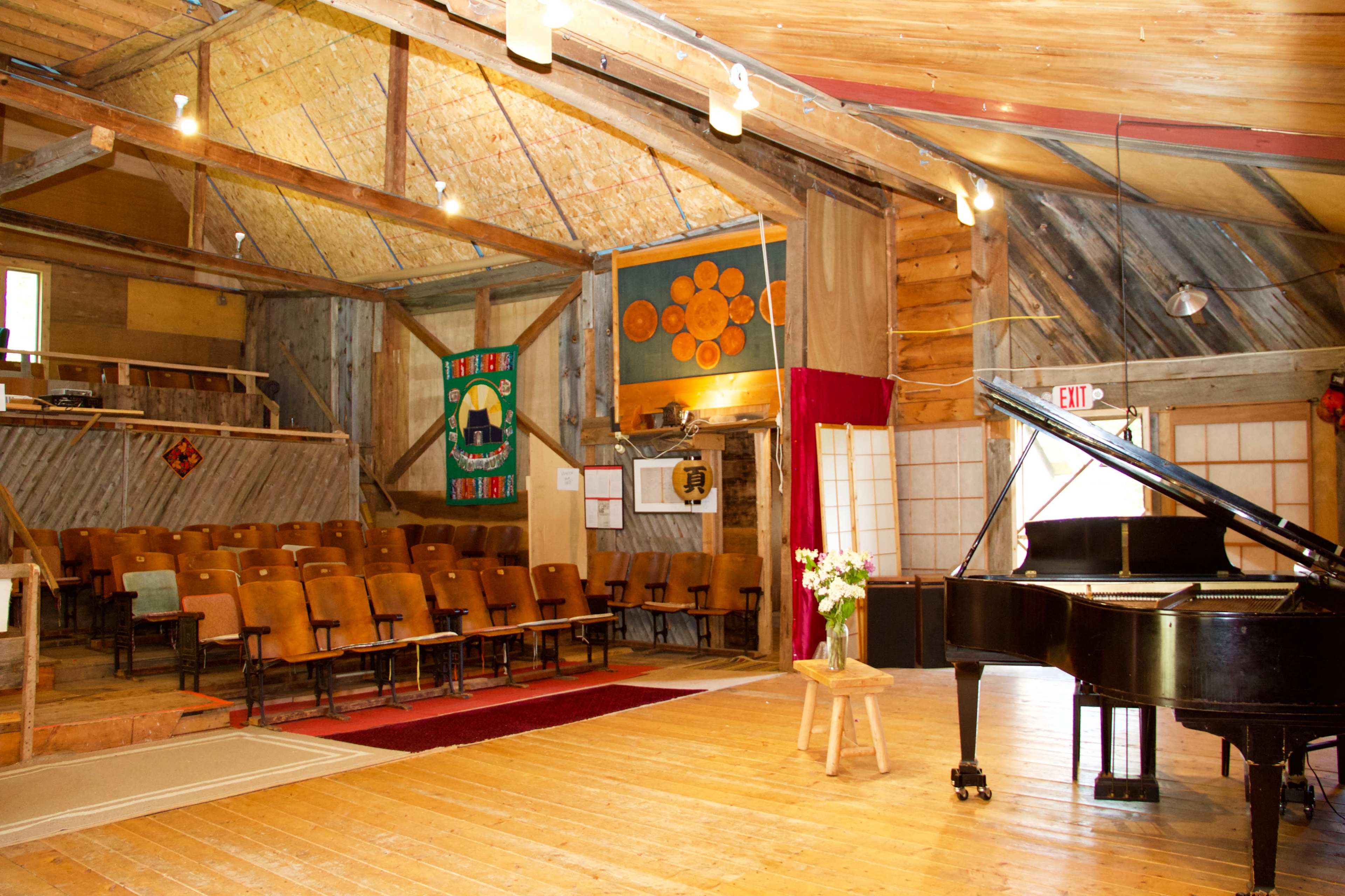 The interior of a rustic hall features a grand piano, wooden floors, rows of orange padded chairs, and colorful banners on the walls.