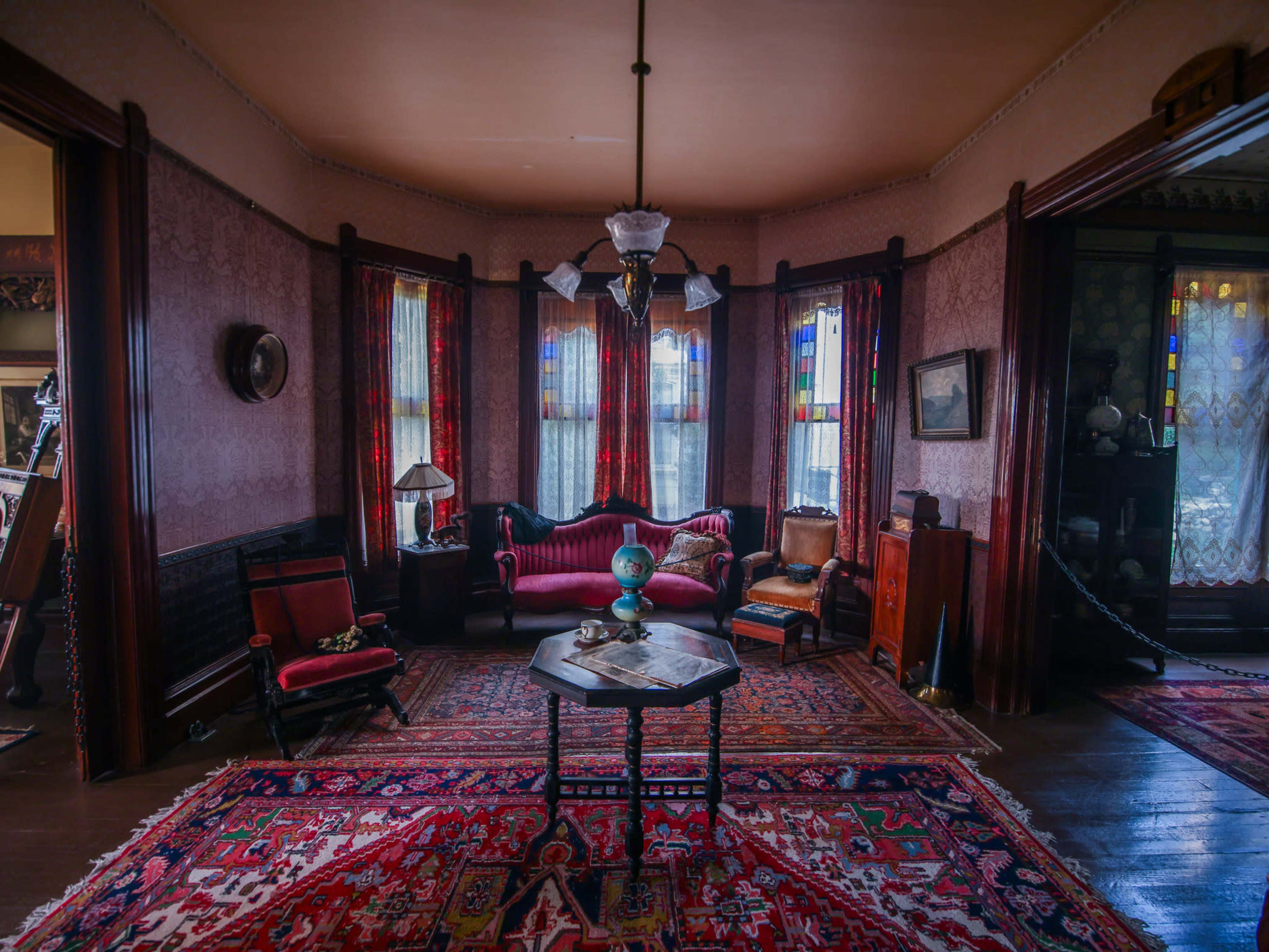 A vintage parlor room with a red patterned rug, a chandelier, stained glass windows, and various pieces of antique furniture.