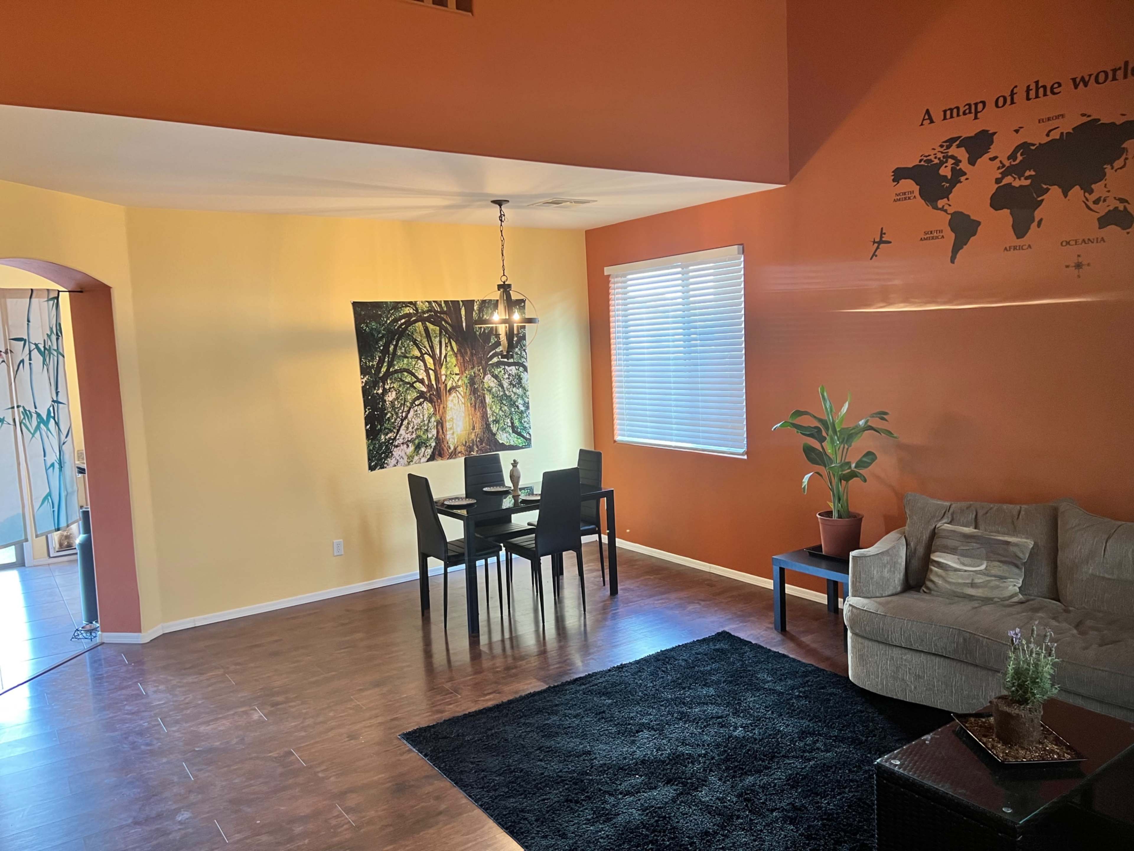 A stylish living space featuring a dining area with a dark table and chairs, a large wall mural of a tree, a cozy couch, and a potted plant.