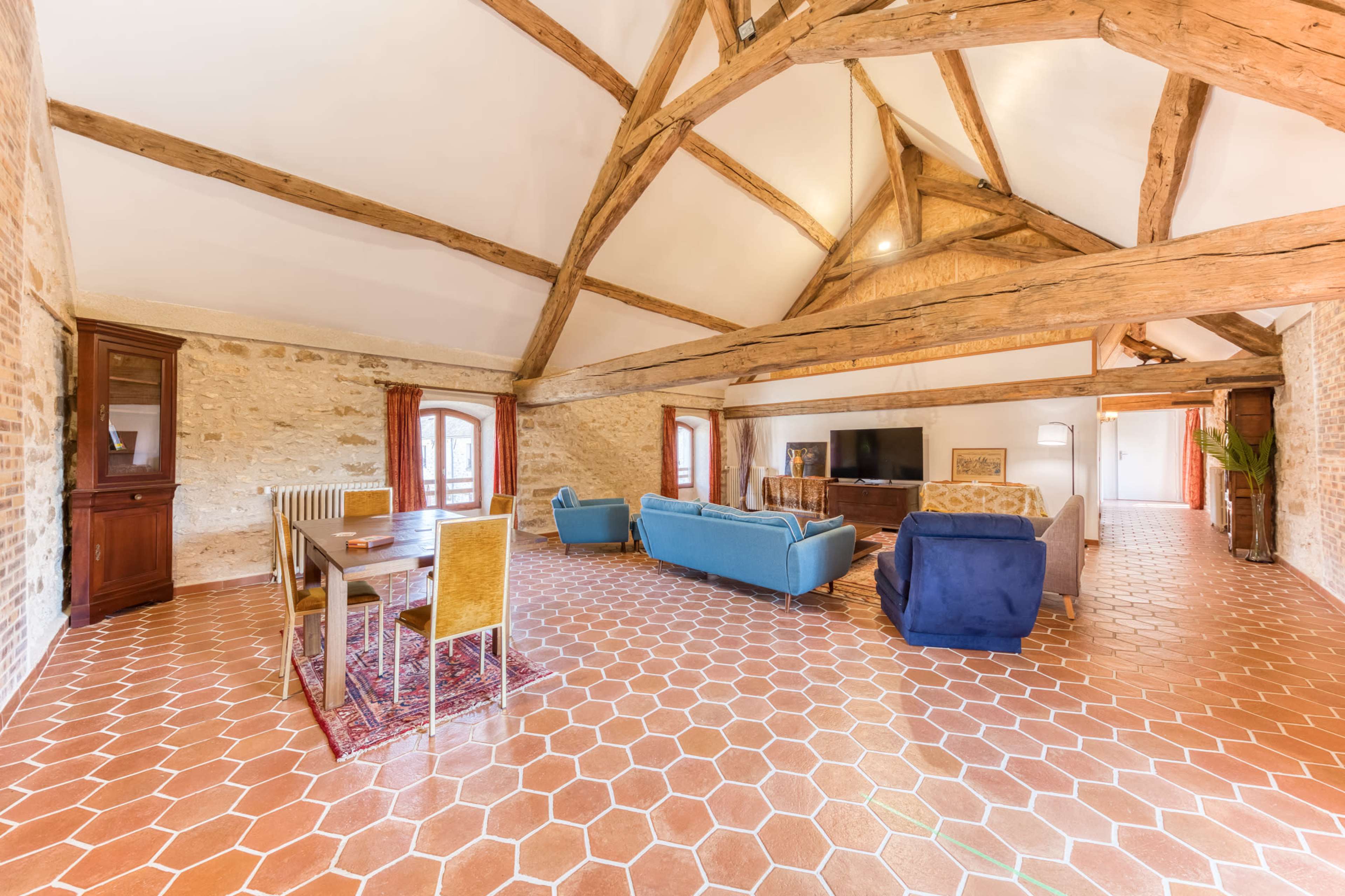 A spacious living area features a mix of rustic brick and wood beam architecture, with a seating arrangement and a dining table set on patterned tiled flooring.