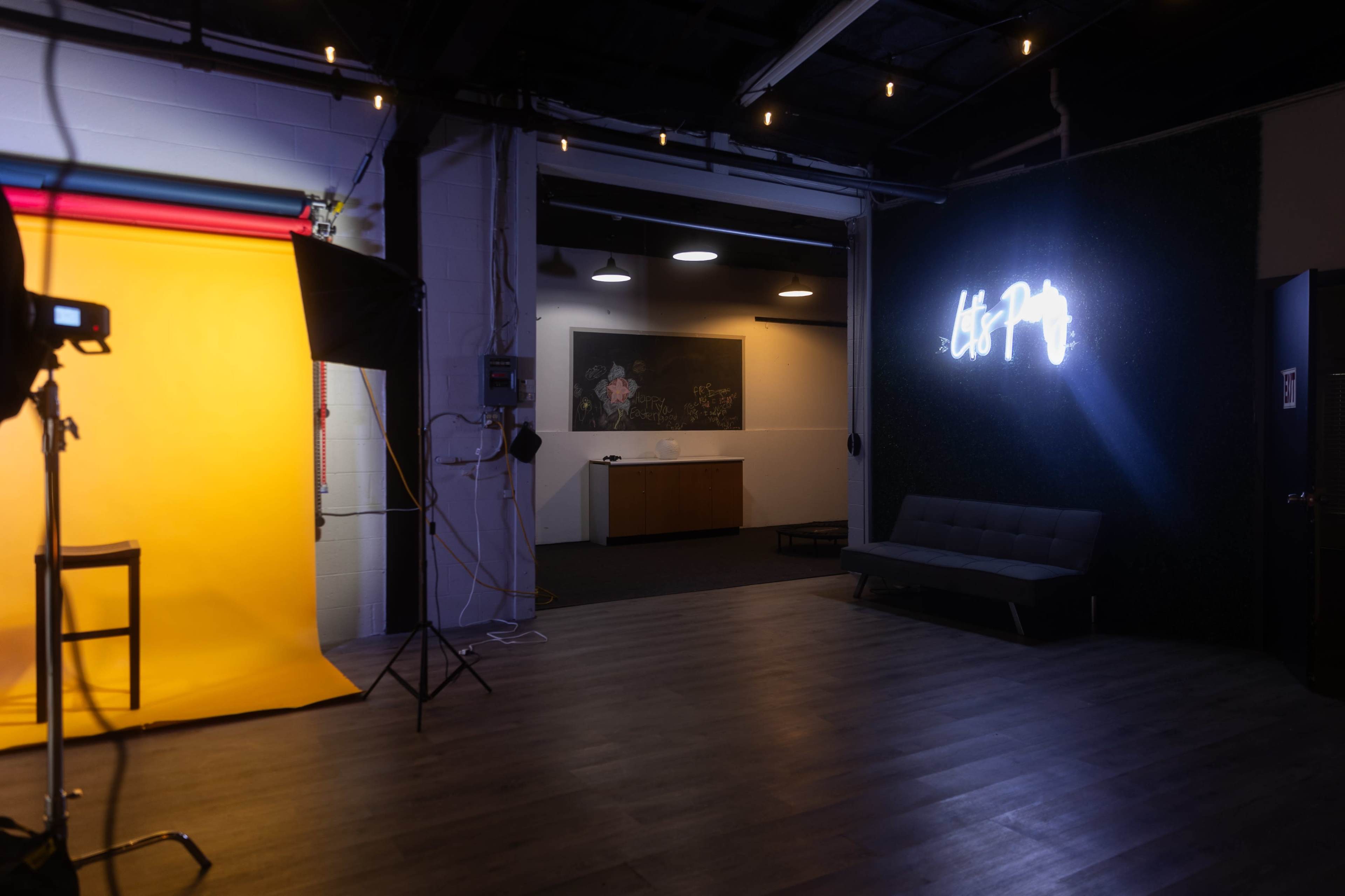 The image shows a modern studio space featuring a yellow backdrop, a camera setup, and a neon sign on a green wall.