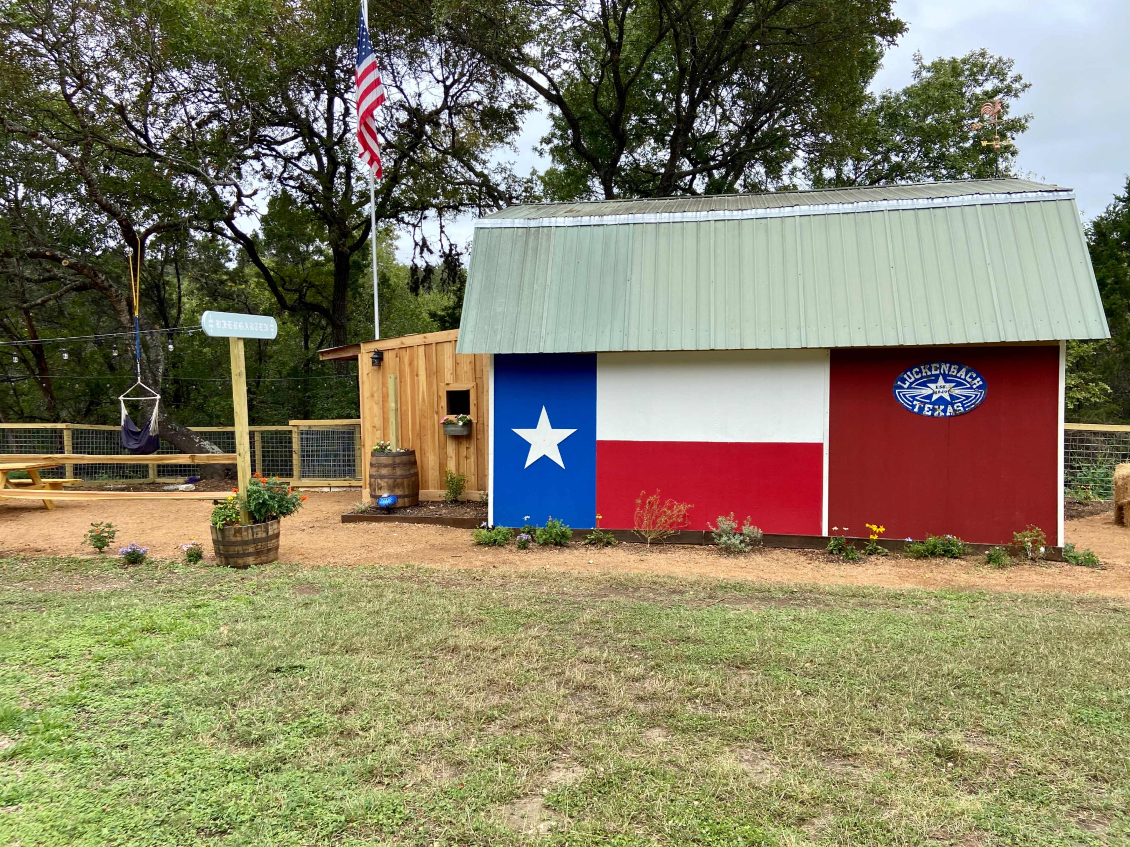 A small shed with a metal roof, featuring a painted Texas flag and a sign that says "Wickerbasket," set in a grassy area surrounded by trees.