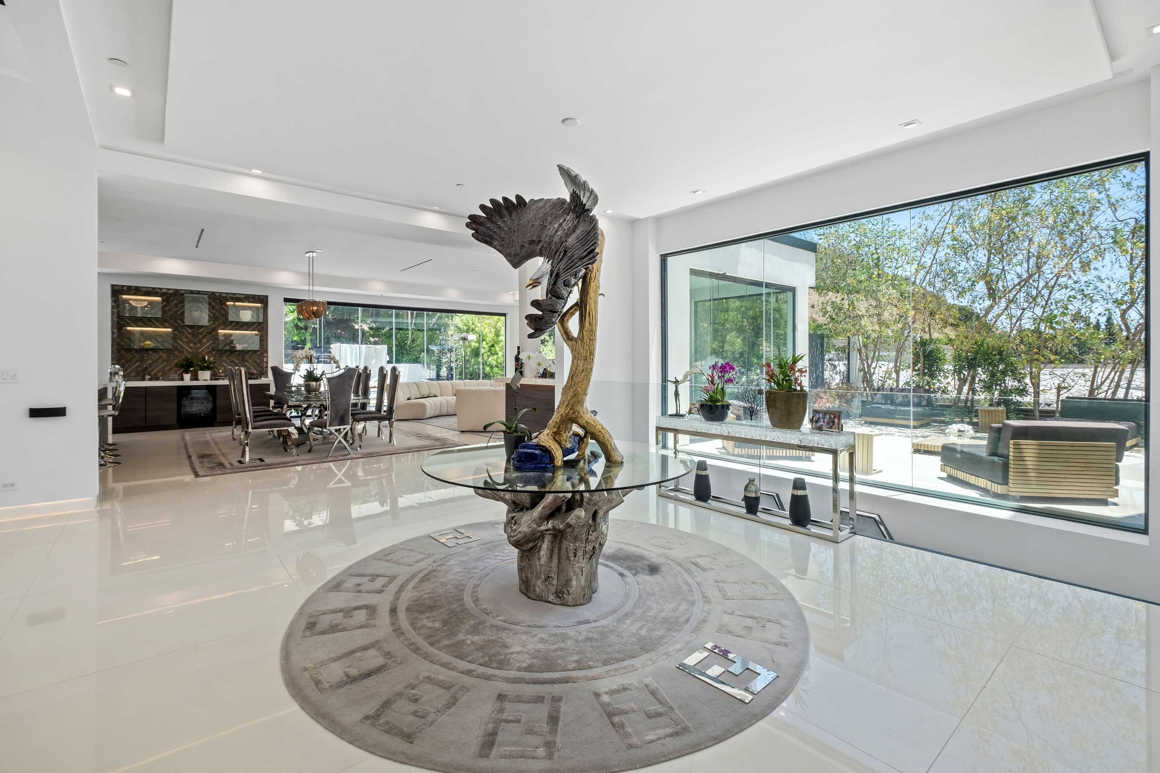 The image depicts a modern living space featuring a glass-topped table with a sculptural base in the center, surrounded by large windows that provide natural light and views of the outdoor area.