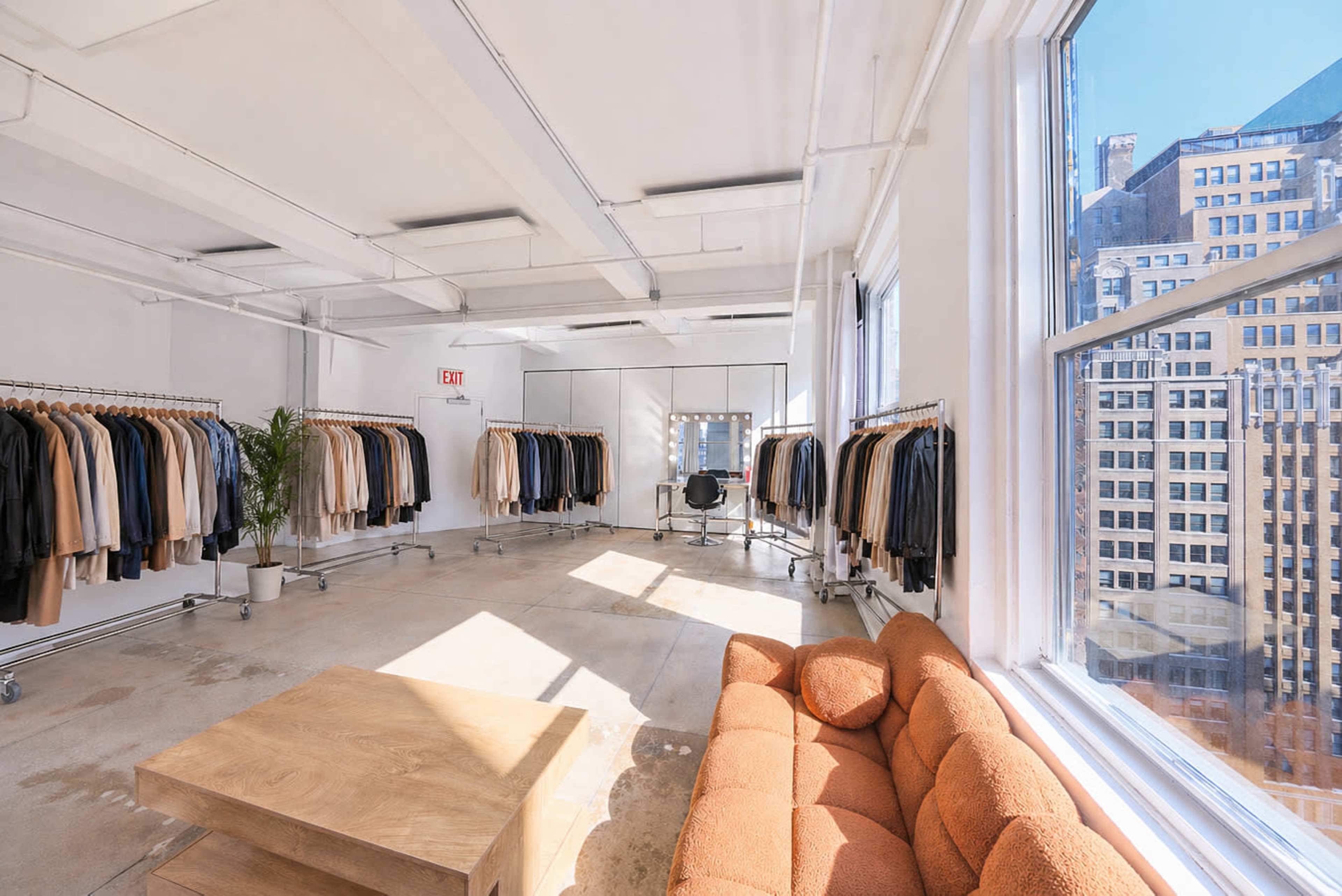 The image shows a well-lit showroom featuring racks of coats on one side and a seating area with a couch and a table near large windows overlooking a cityscape.