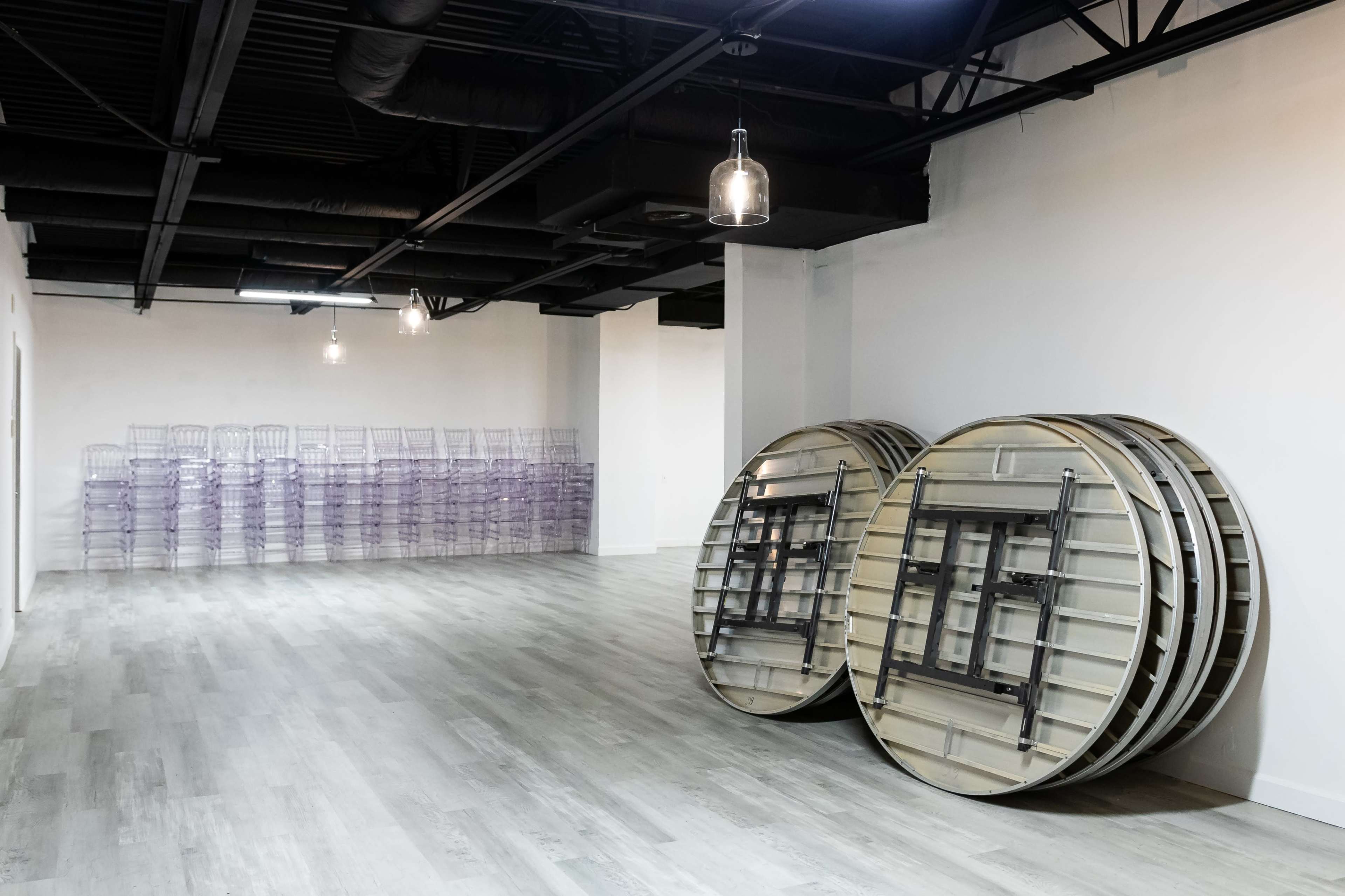 The image shows an empty room with two stacked round tables and a row of transparent chairs stacked against the wall.
