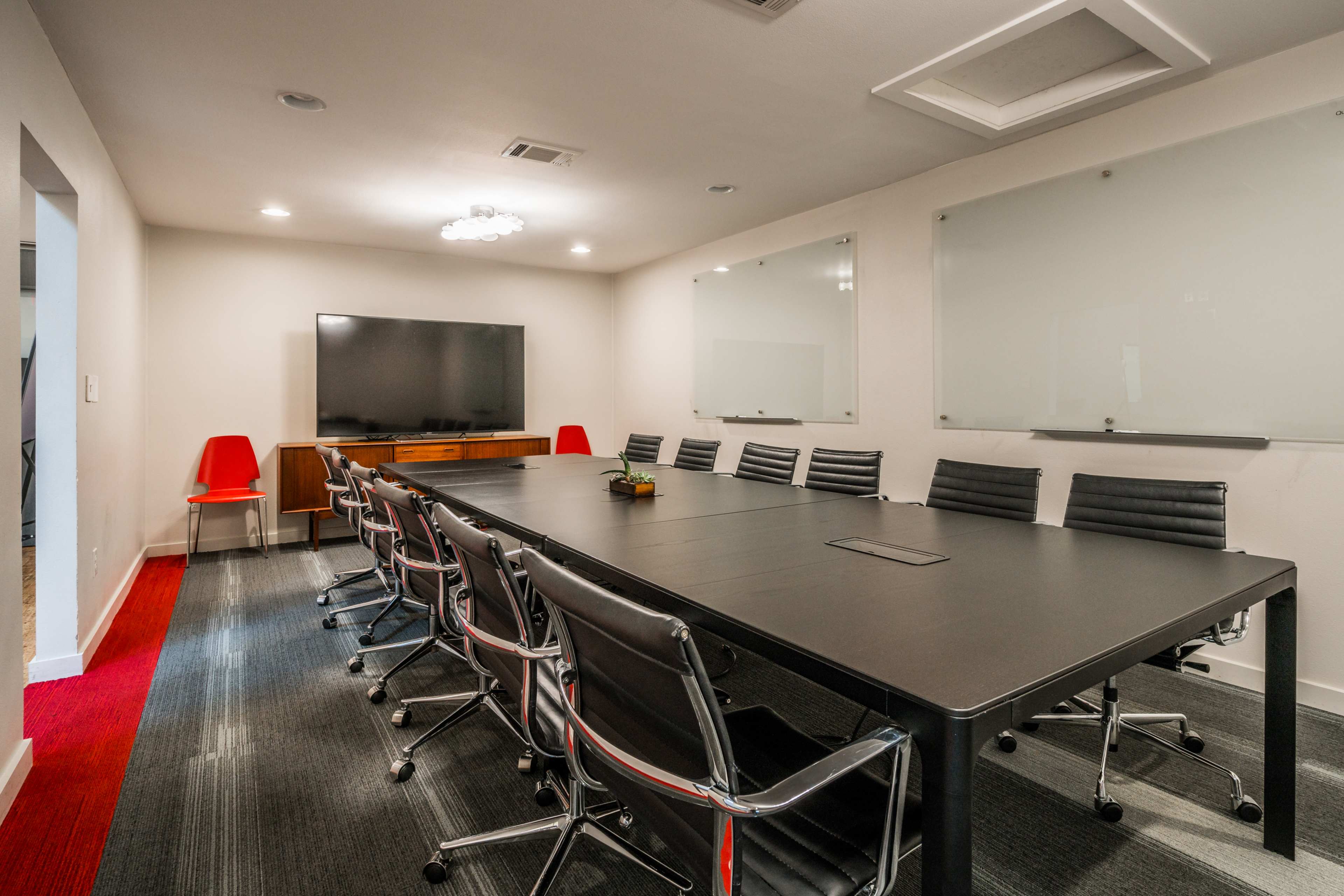 A modern conference room with a long black table, black rolling chairs, a large television screen on one wall, and whiteboard panels on the opposite wall.