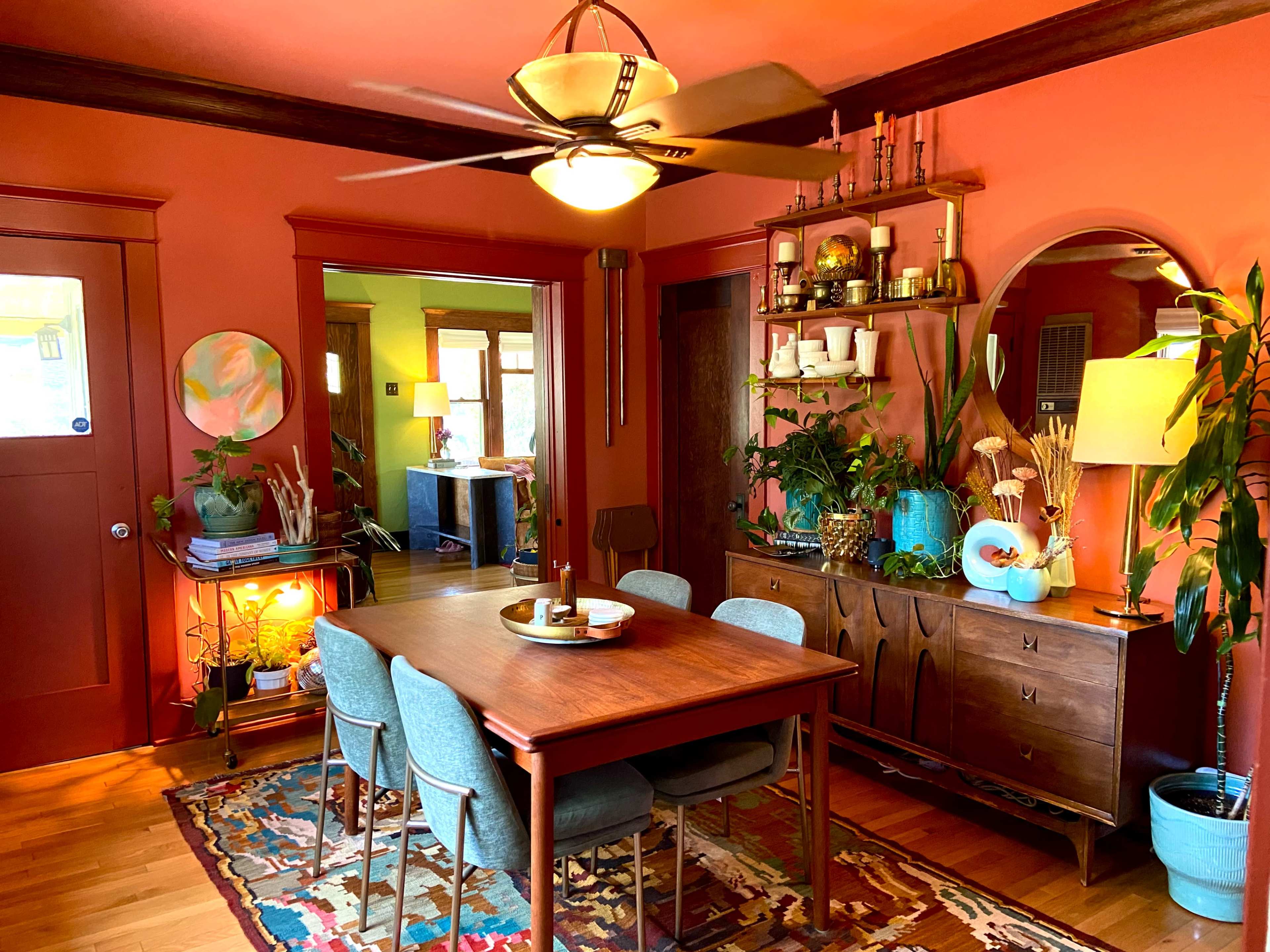 The image shows a cozy dining room with an orange wall, a wooden table surrounded by blue chairs, a ceiling fan, and various decorative plants and accessories.
