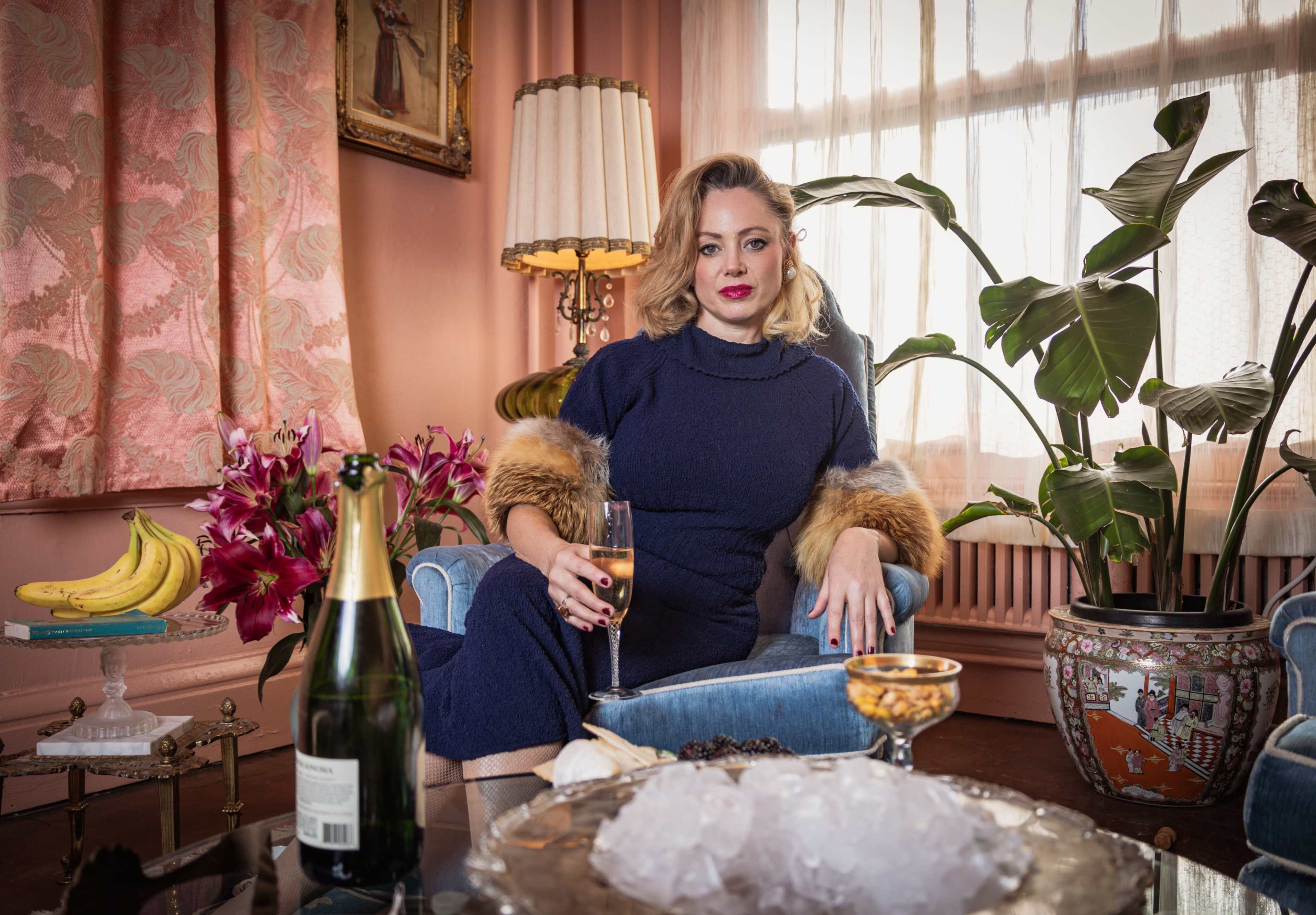 A woman in a navy dress sits elegantly on a blue chair in a vintage-styled room adorned with plants and flowers, holding a glass of champagne.