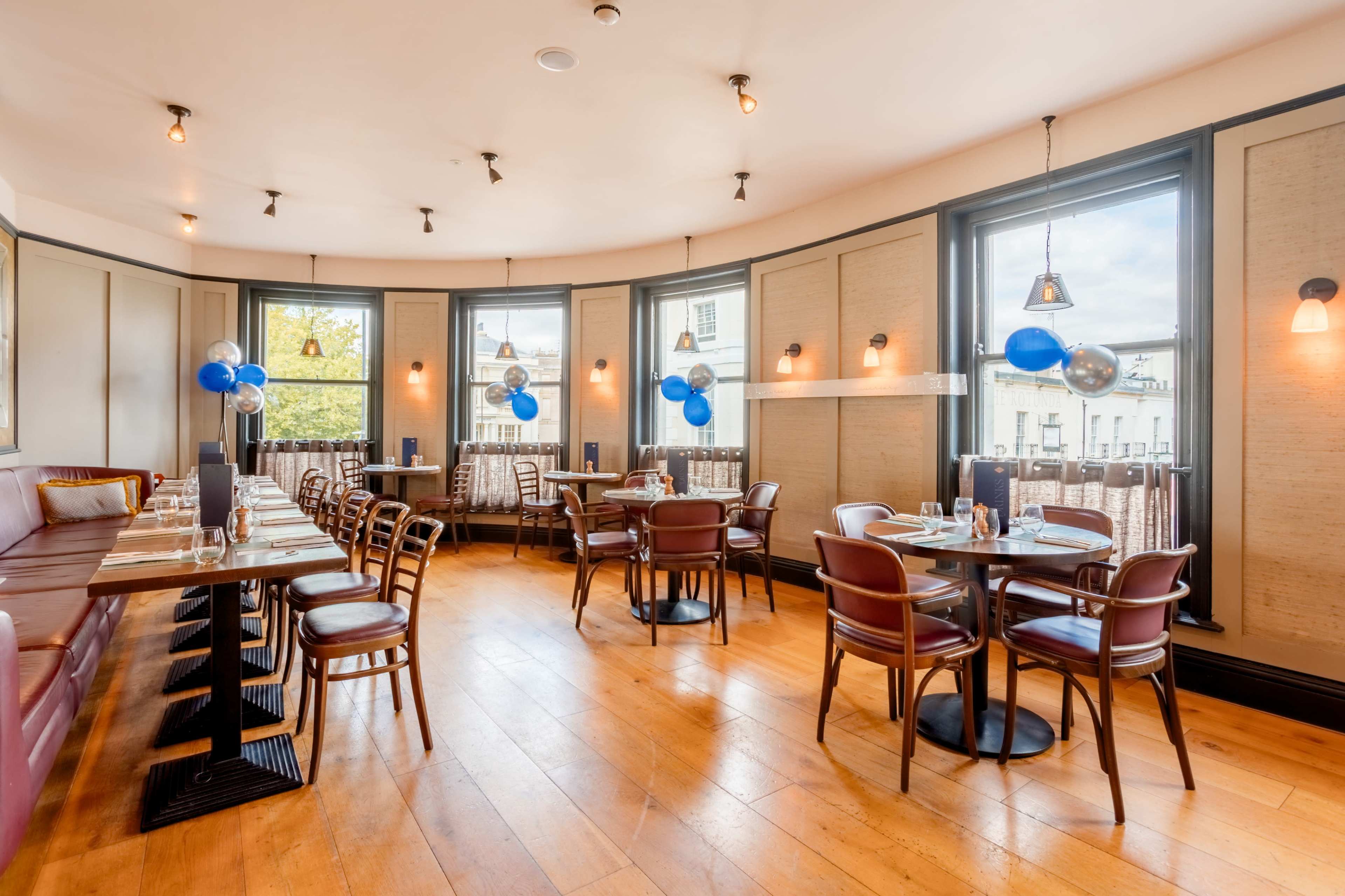 The image shows a modern restaurant interior with wooden floors, brown chairs, and large windows, decorated with blue balloons.