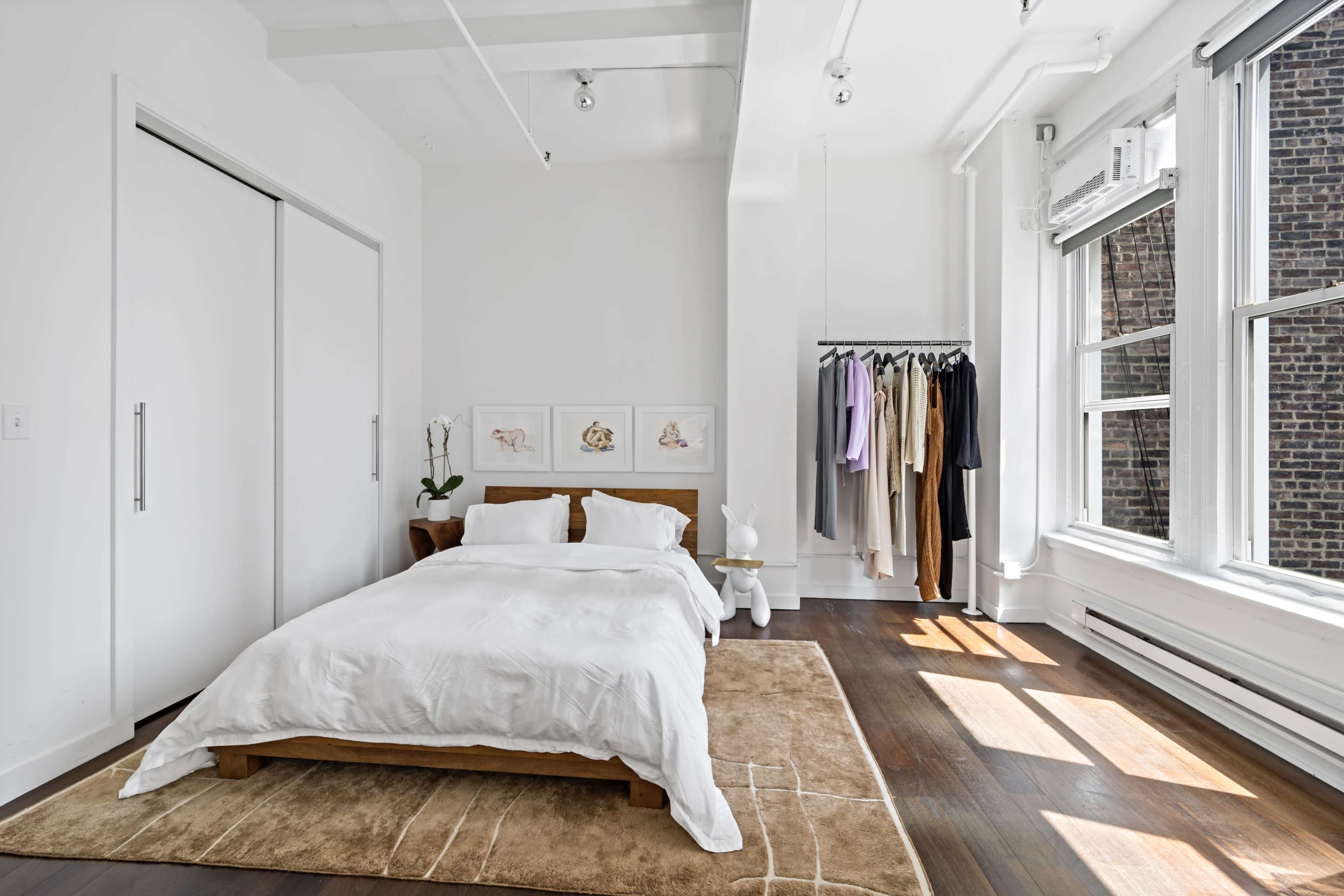 A minimalist bedroom with a wooden bed, a neutral area rug, and a clothing rack displaying various garments near large windows.