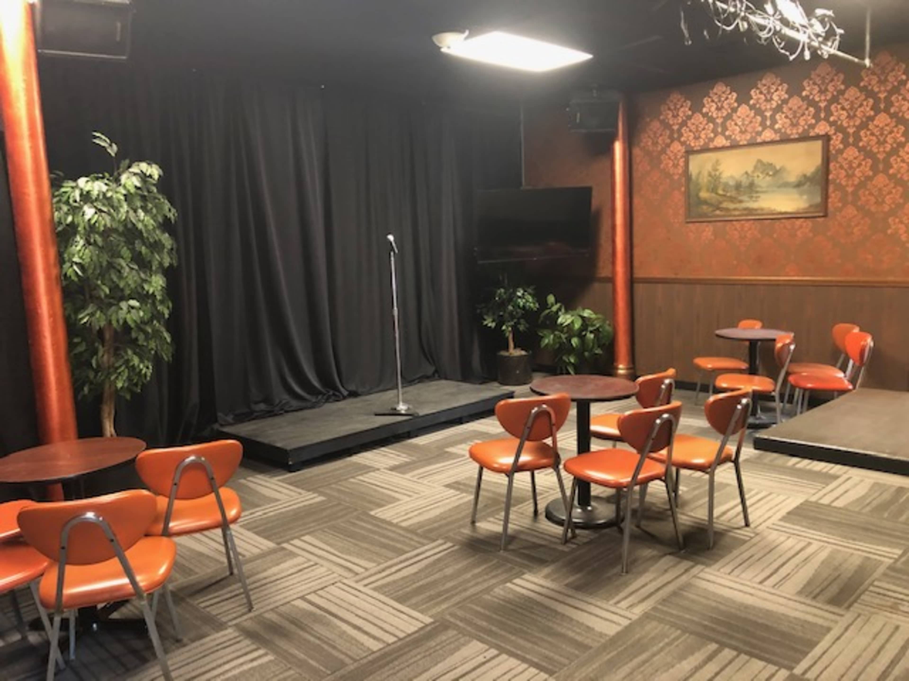 The image shows a small performance stage with a microphone, surrounded by several orange chairs and small tables in a dimly lit room with a decorative backdrop.