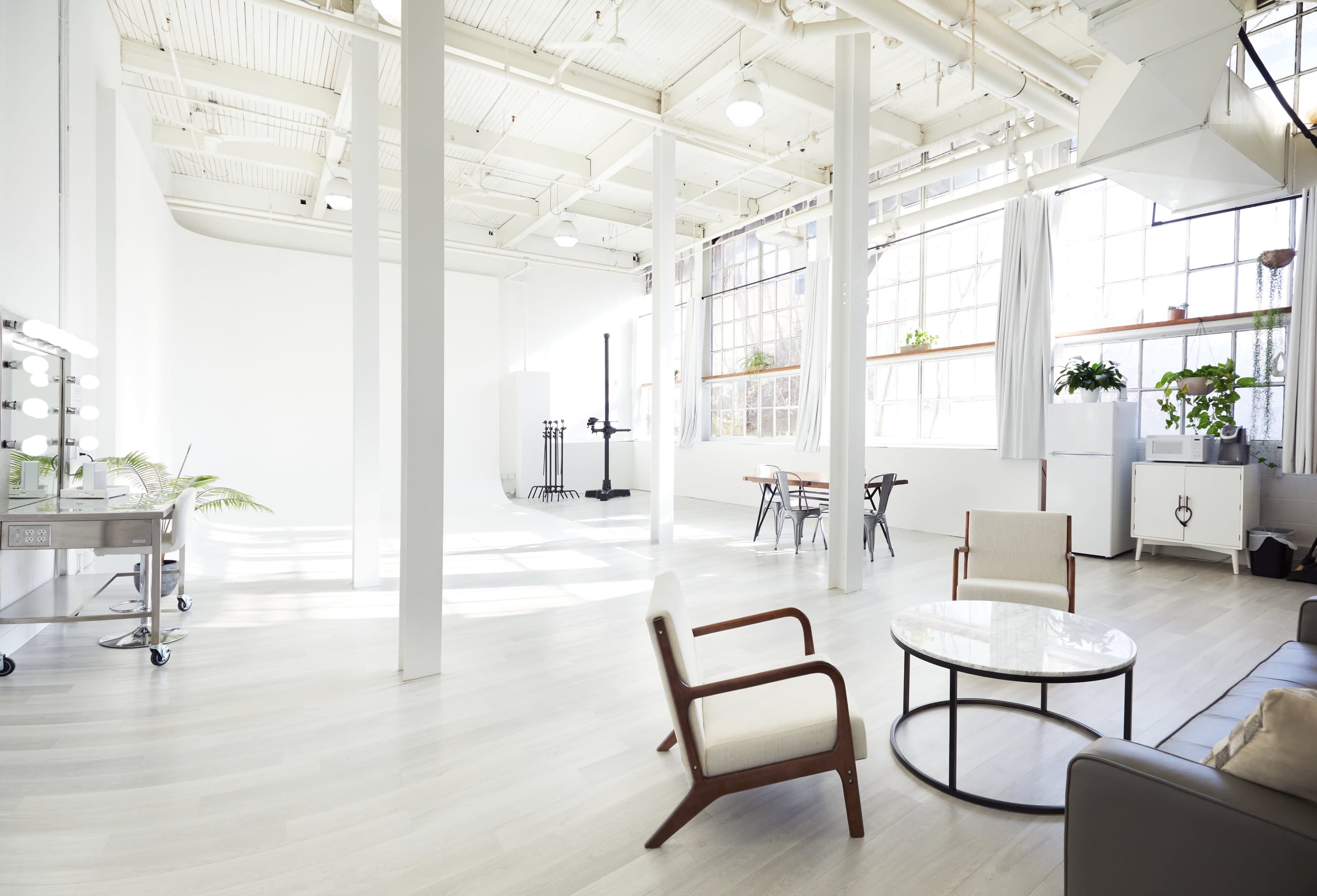 The image shows a spacious, well-lit room with white walls, large windows, and minimalist furniture, including a round coffee table, chairs, and a dining area.