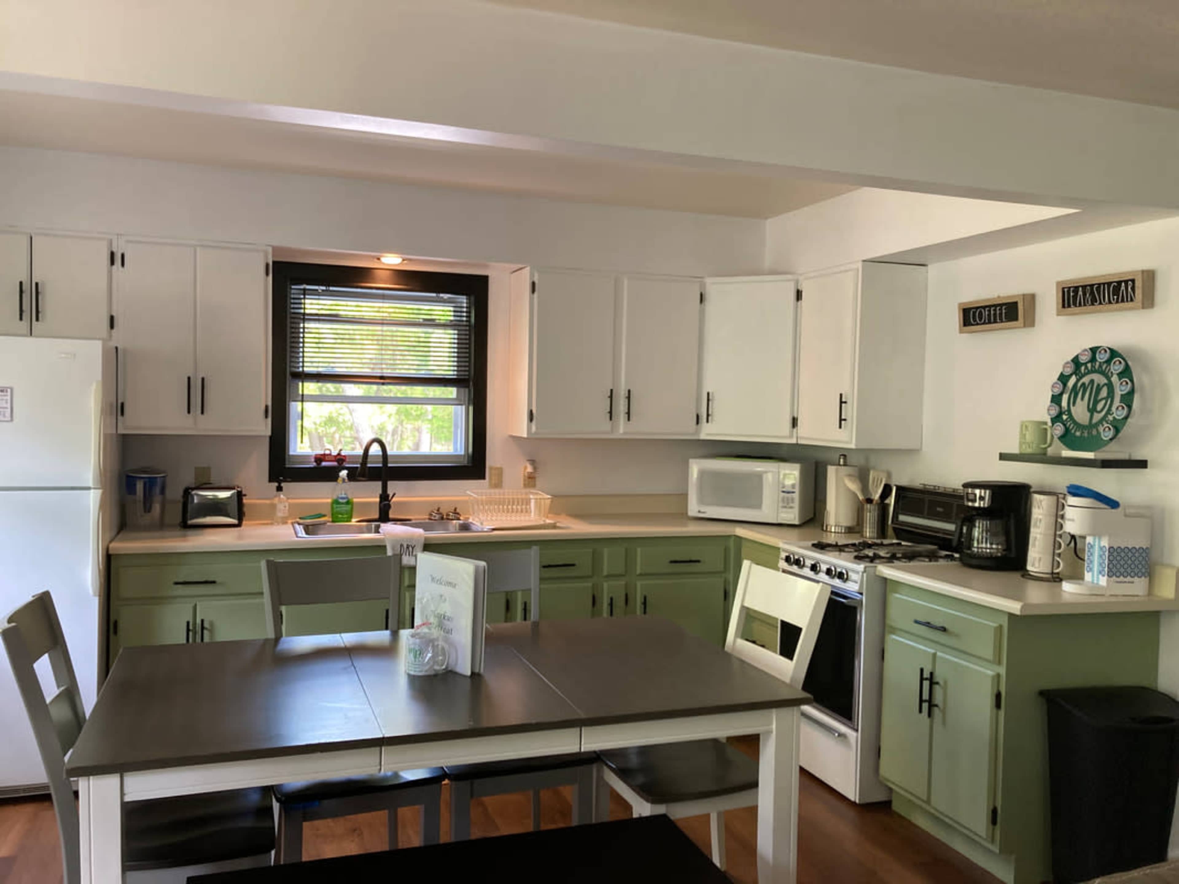 A modern kitchen features white cabinets with green accents, a countertop with appliances, and a dining table set for four.