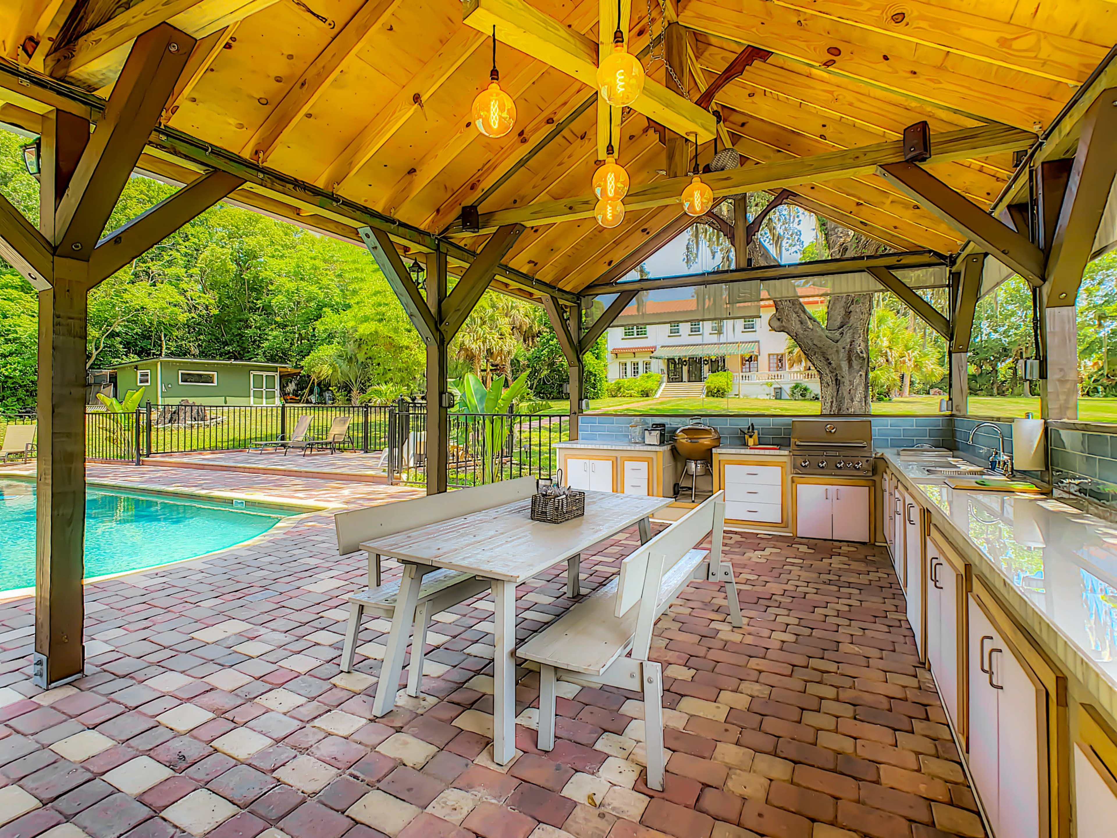 An outdoor kitchen area with a wooden pavilion, a large dining table, and a swimming pool visible in the background.