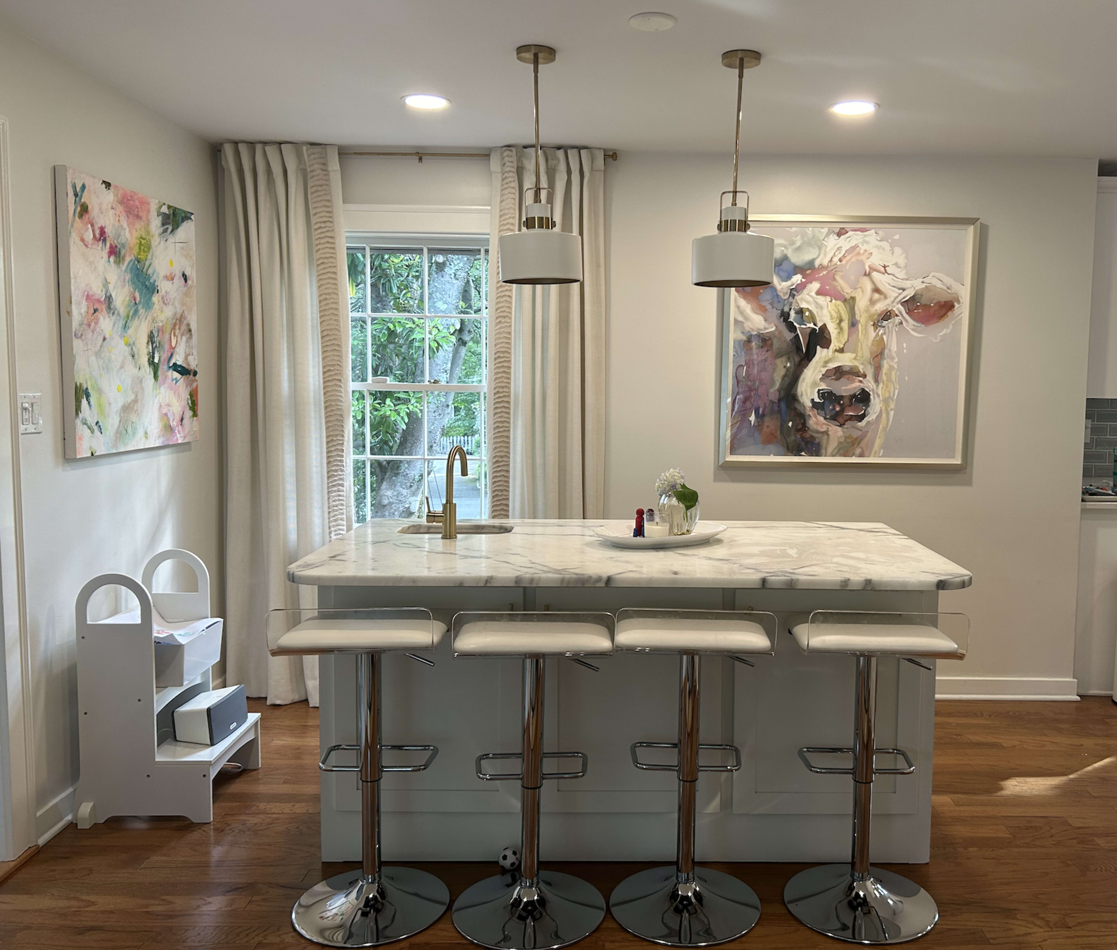 A modern kitchen features a marble island with four stools, large windows, and artwork on the walls.