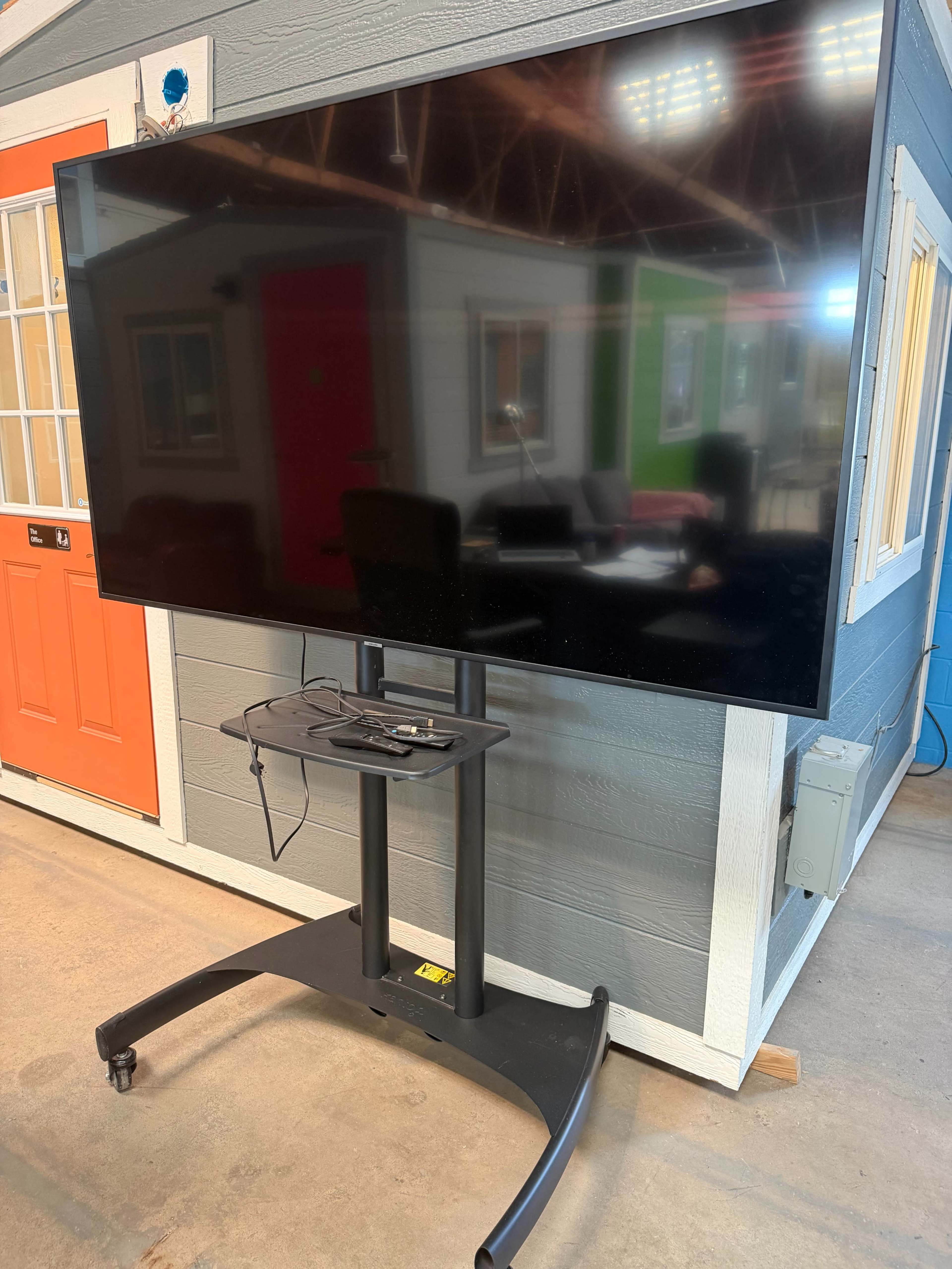 The image shows a large flat-screen television mounted on a wheeled stand in a brightly colored room with a desk and chairs visible in the background.