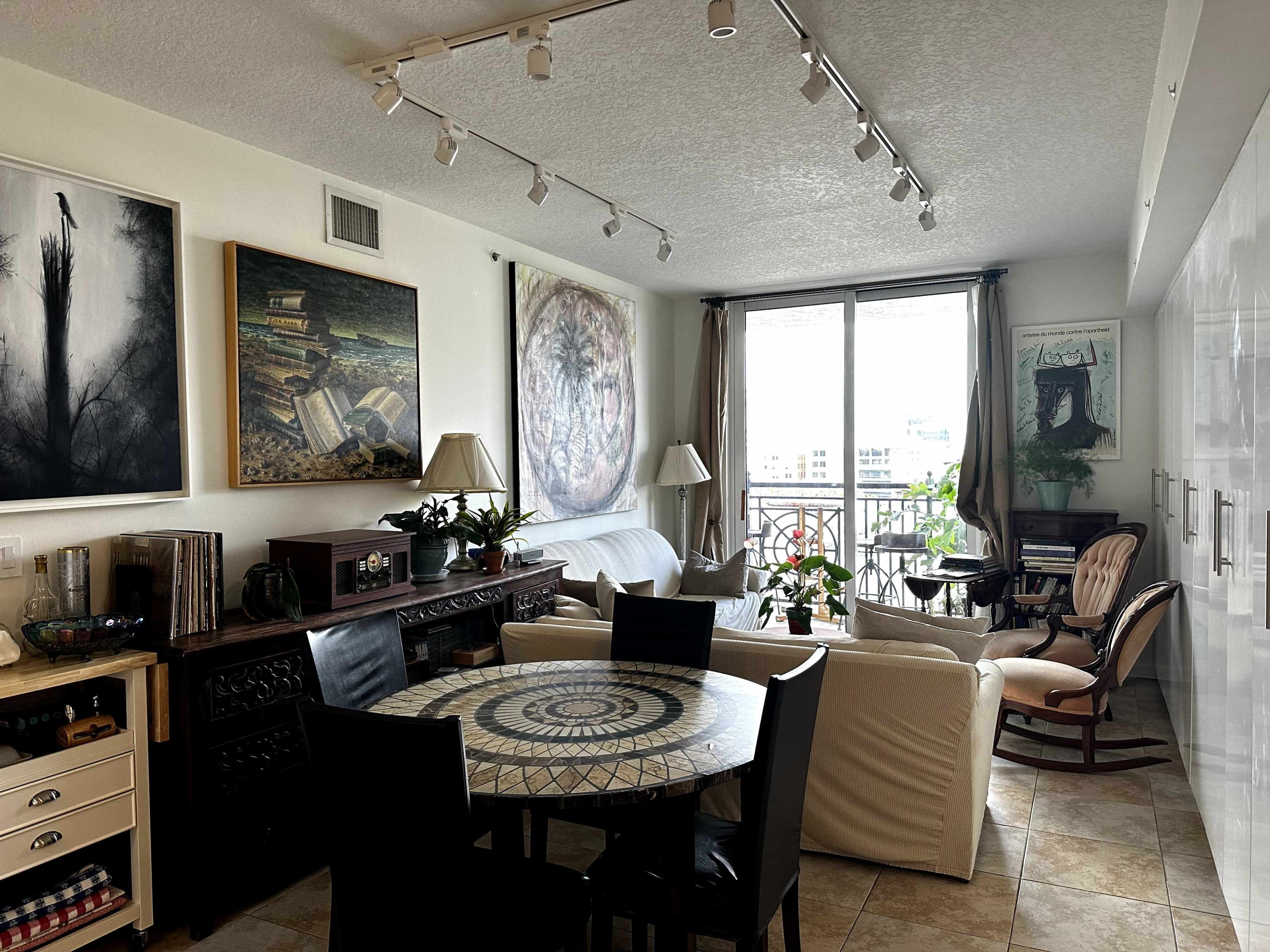 A modern living room with a circular patterned dining table, a sofa, and various artworks on the walls, along with large windows letting in natural light.