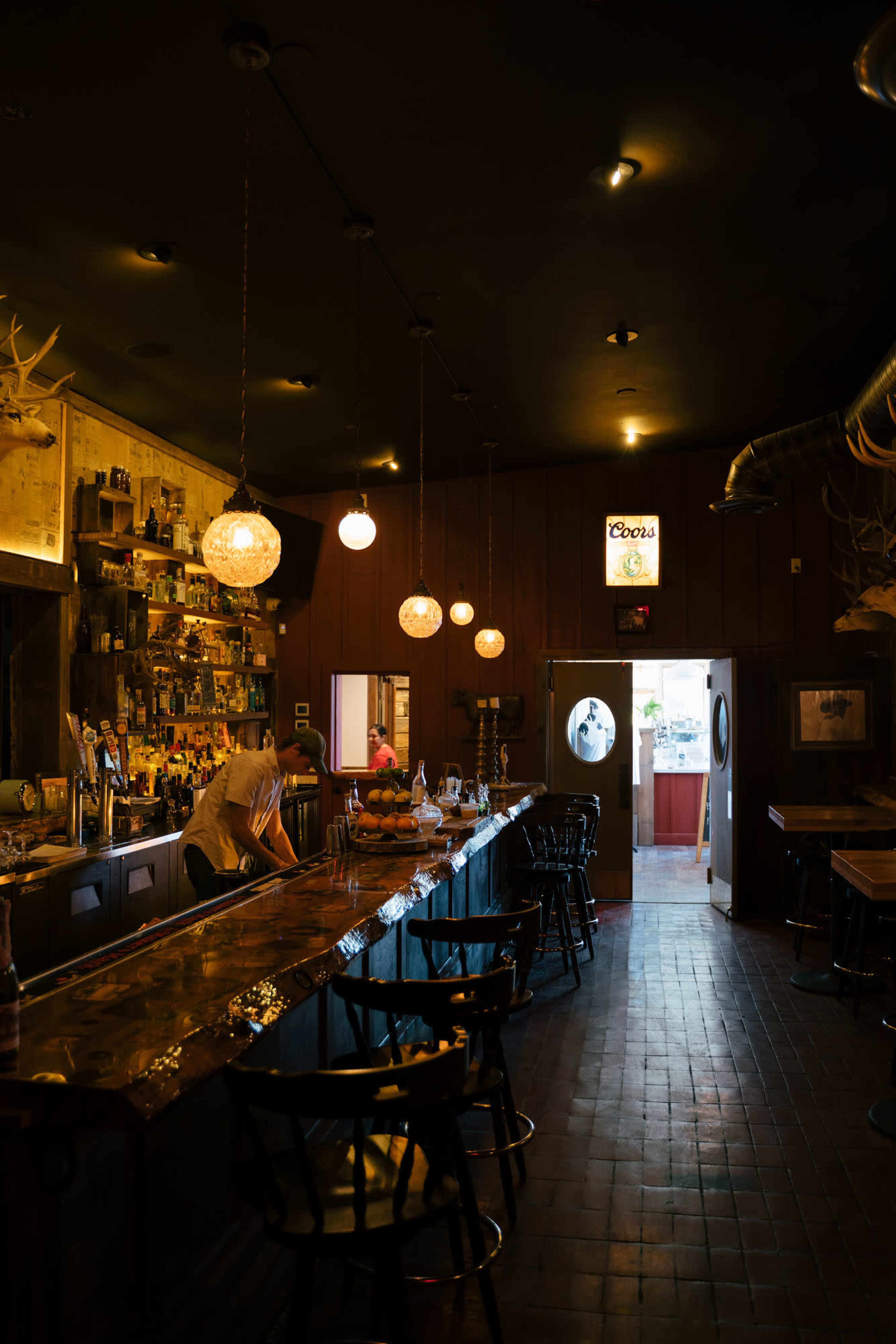 A dimly lit bar features a long counter with several stools, shelves lined with bottles, and round pendant lights hanging from the ceiling.