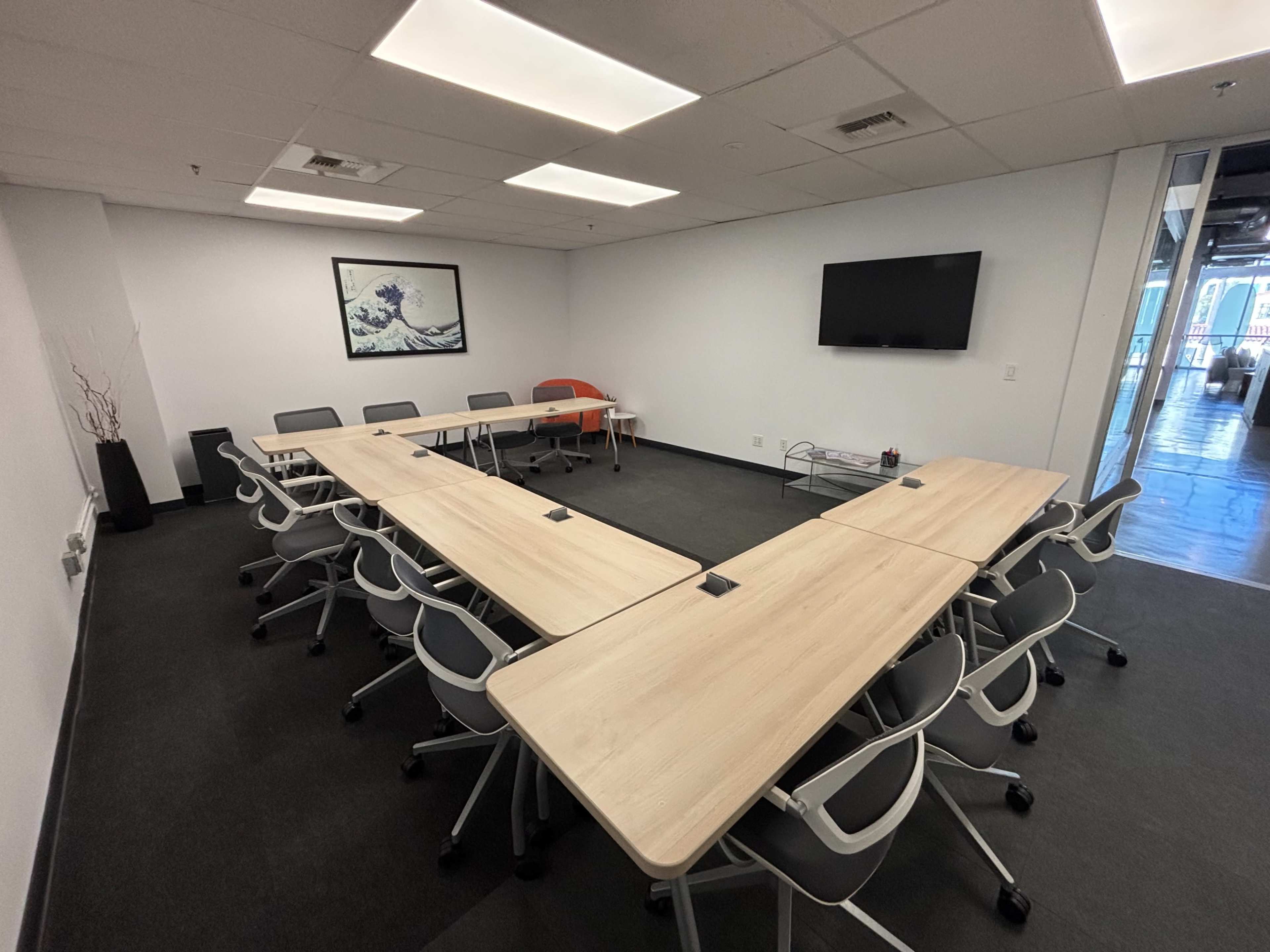 The image shows a modern conference room with several rectangular tables arranged in a U-shape, equipped with chairs and a wall-mounted television.