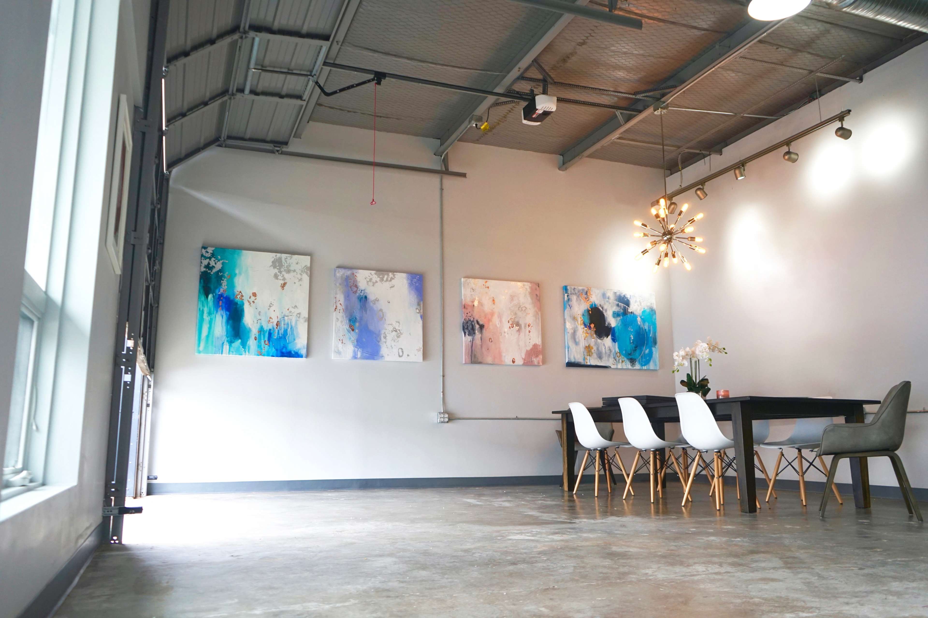 A modern dining area features a long table with stylish chairs, illuminated by a contemporary light fixture, and is adorned with abstract paintings on the walls.