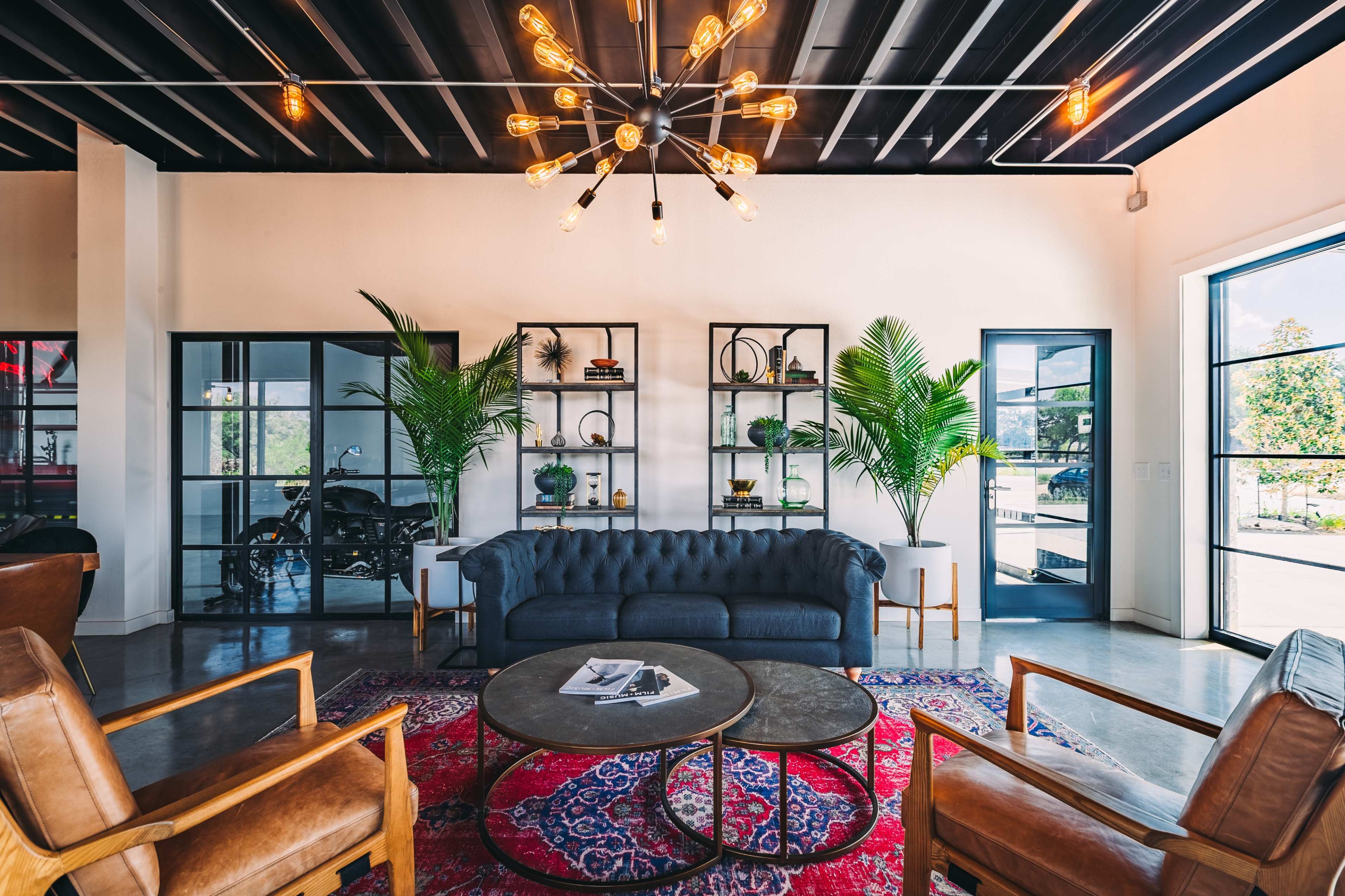 The scene shows a modern lounge area featuring a tufted sofa, wooden chairs, a round coffee table, and open shelves with plants, all under a decorative ceiling light.