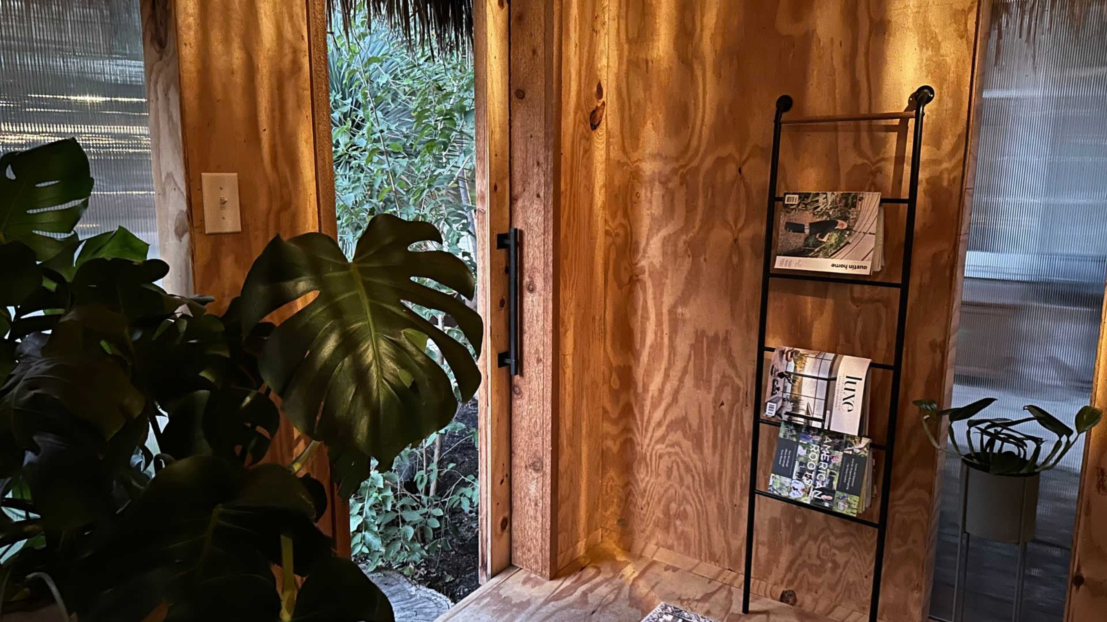 The image shows a wooden interior with an open door, a plant in the foreground, and a metal rack holding magazines against the wall.