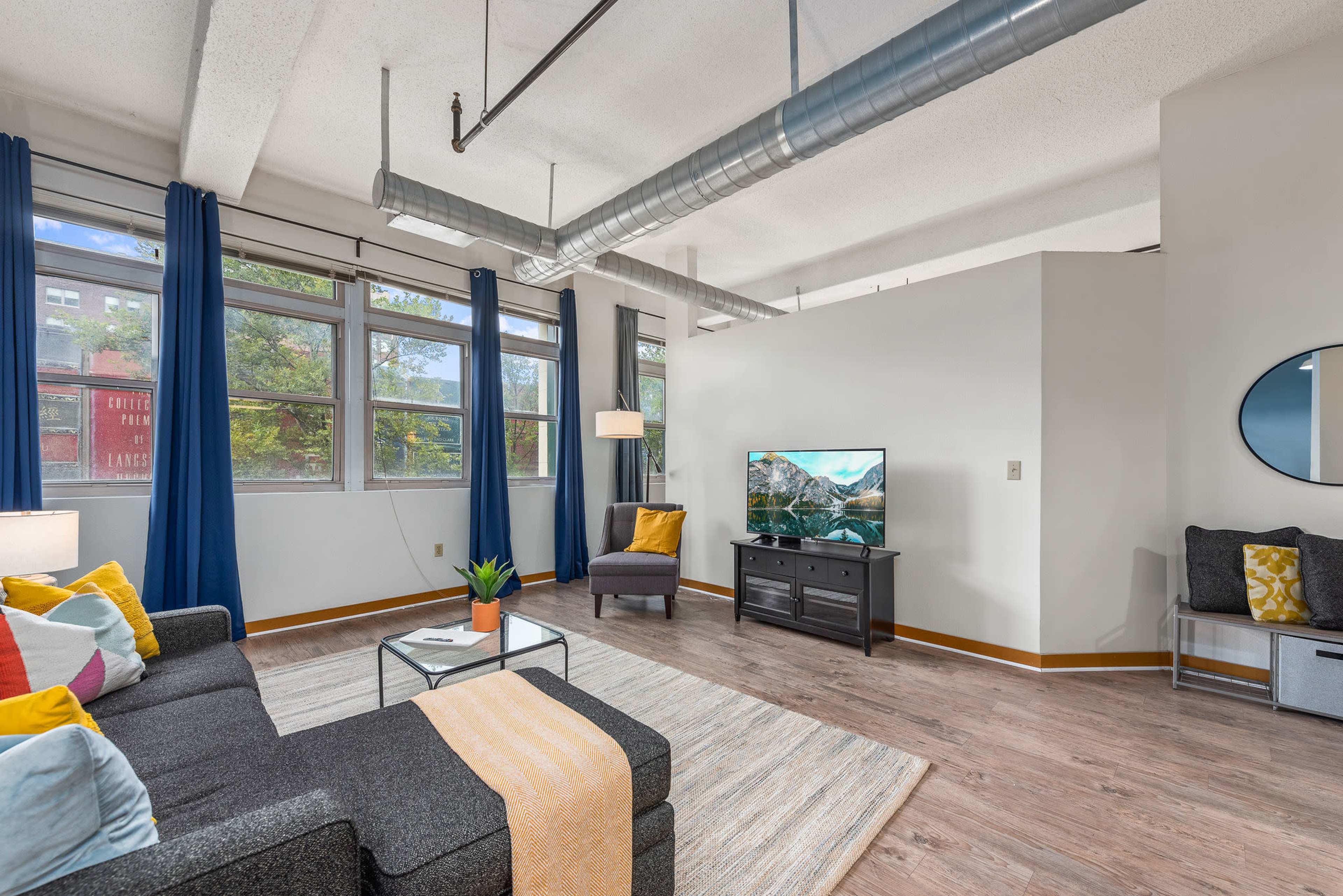 A modern living room features a gray sofa with colorful cushions, a coffee table, a television on a black stand, and large windows with blue curtains.