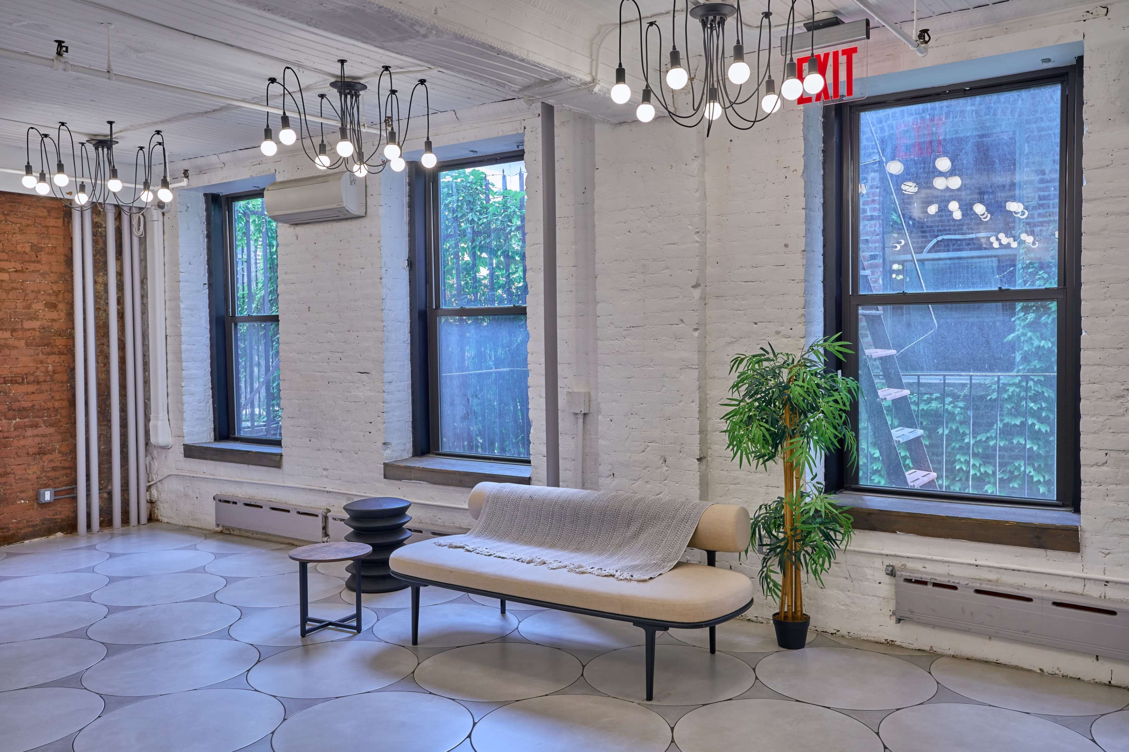 A bright room with large windows, a light-colored sofa, a small black table, and decorative plants, all beneath modern ceiling lights.