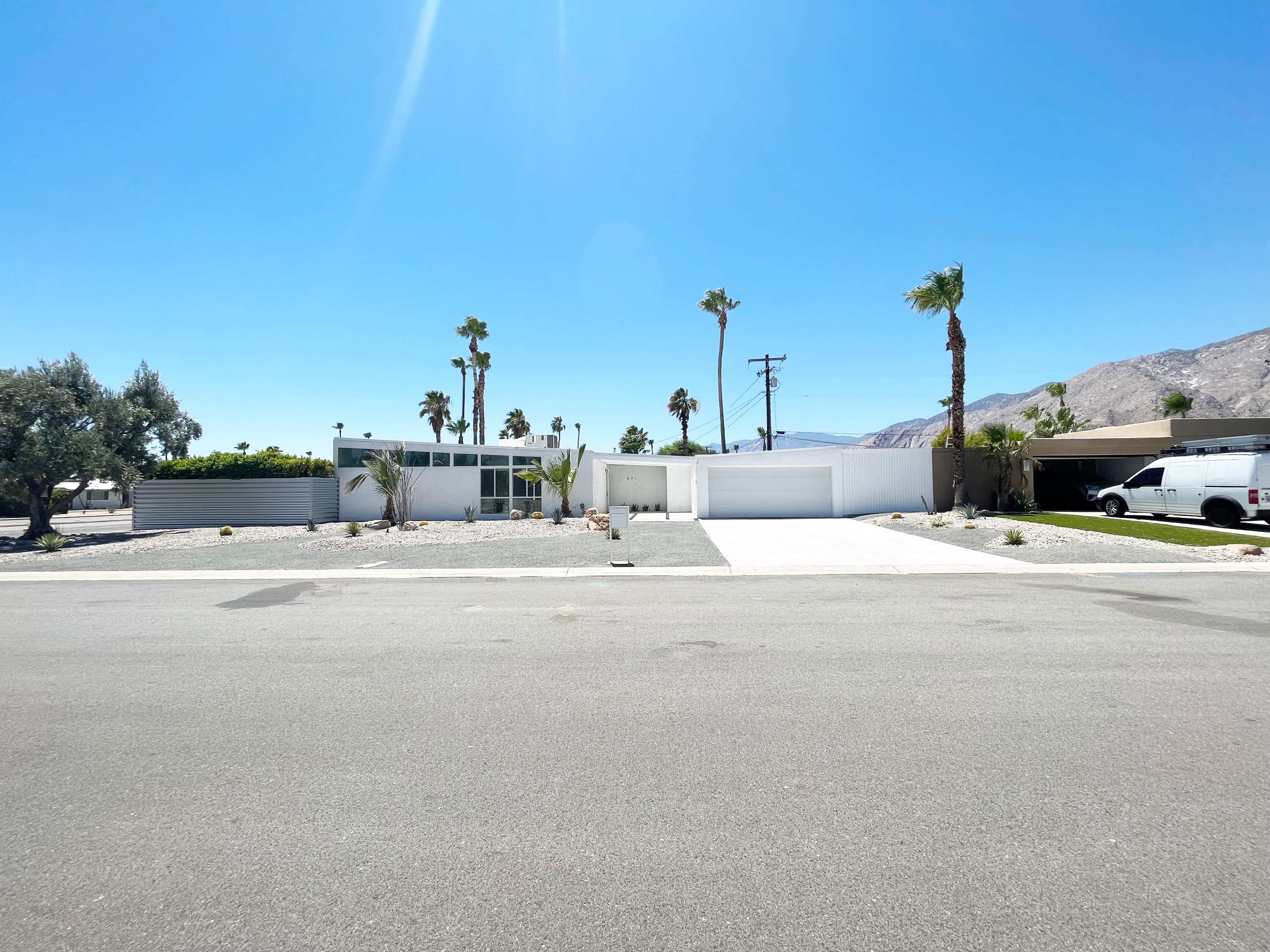 The image shows a modern single-story house with a flat roof, surrounded by palm trees and gravel landscaping, located on a paved street.