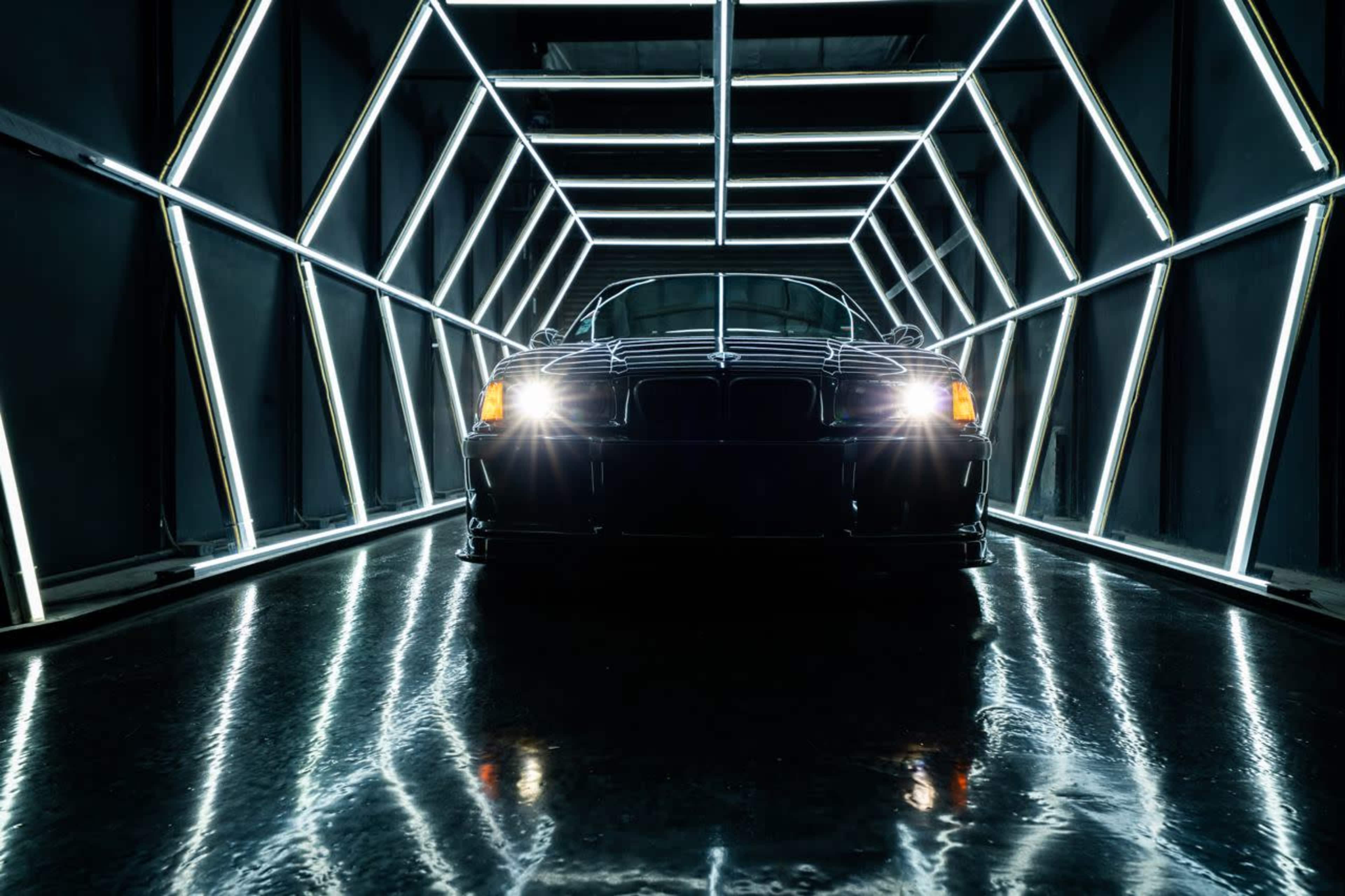 A black car is positioned in a geometric tunnel illuminated by bright, linear neon lights.