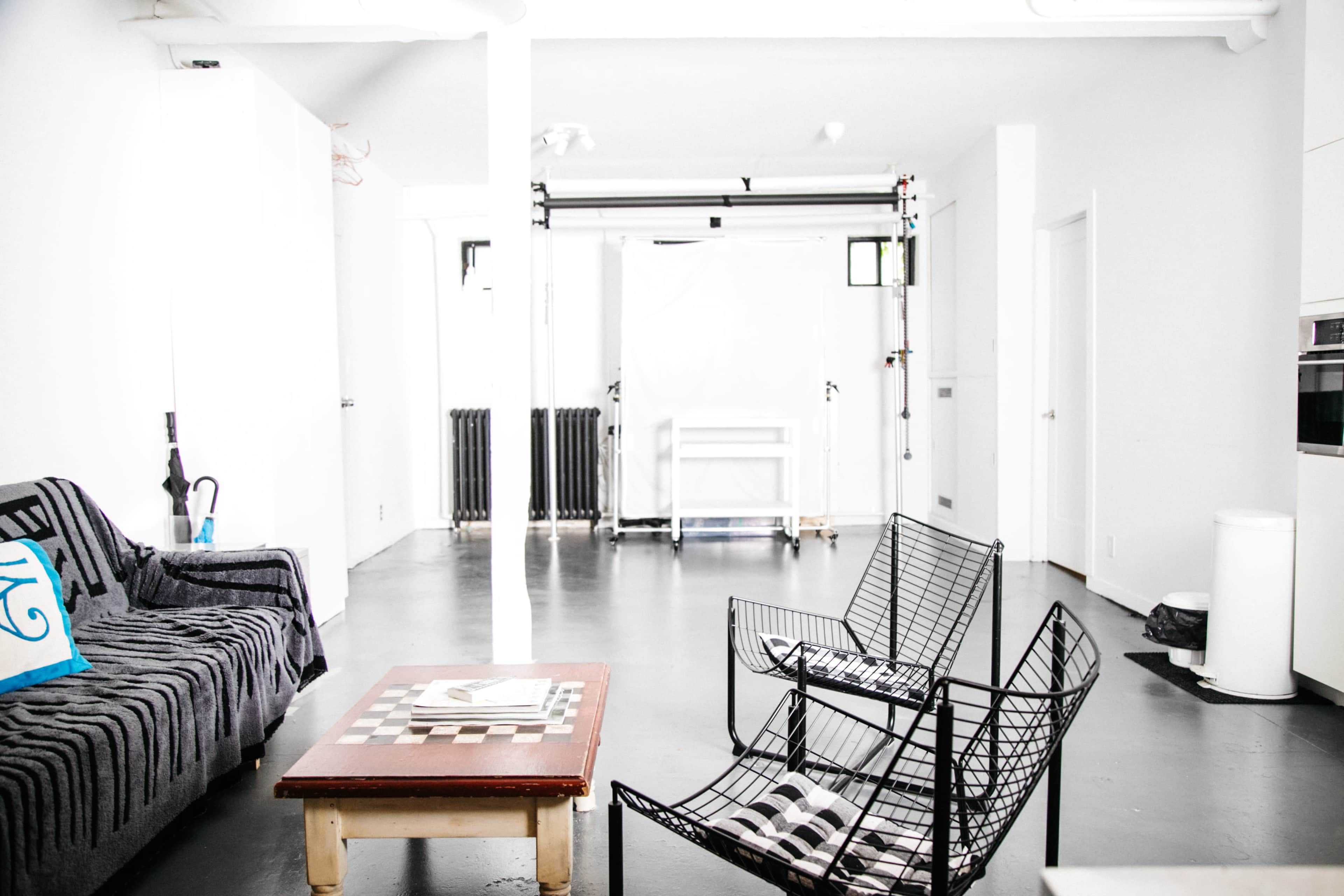 The image shows a minimalist, modern interior featuring a black and white striped sofa, two wireframe chairs, a wooden coffee table with a checkerboard, and a backdrop setup in a bright, white room.