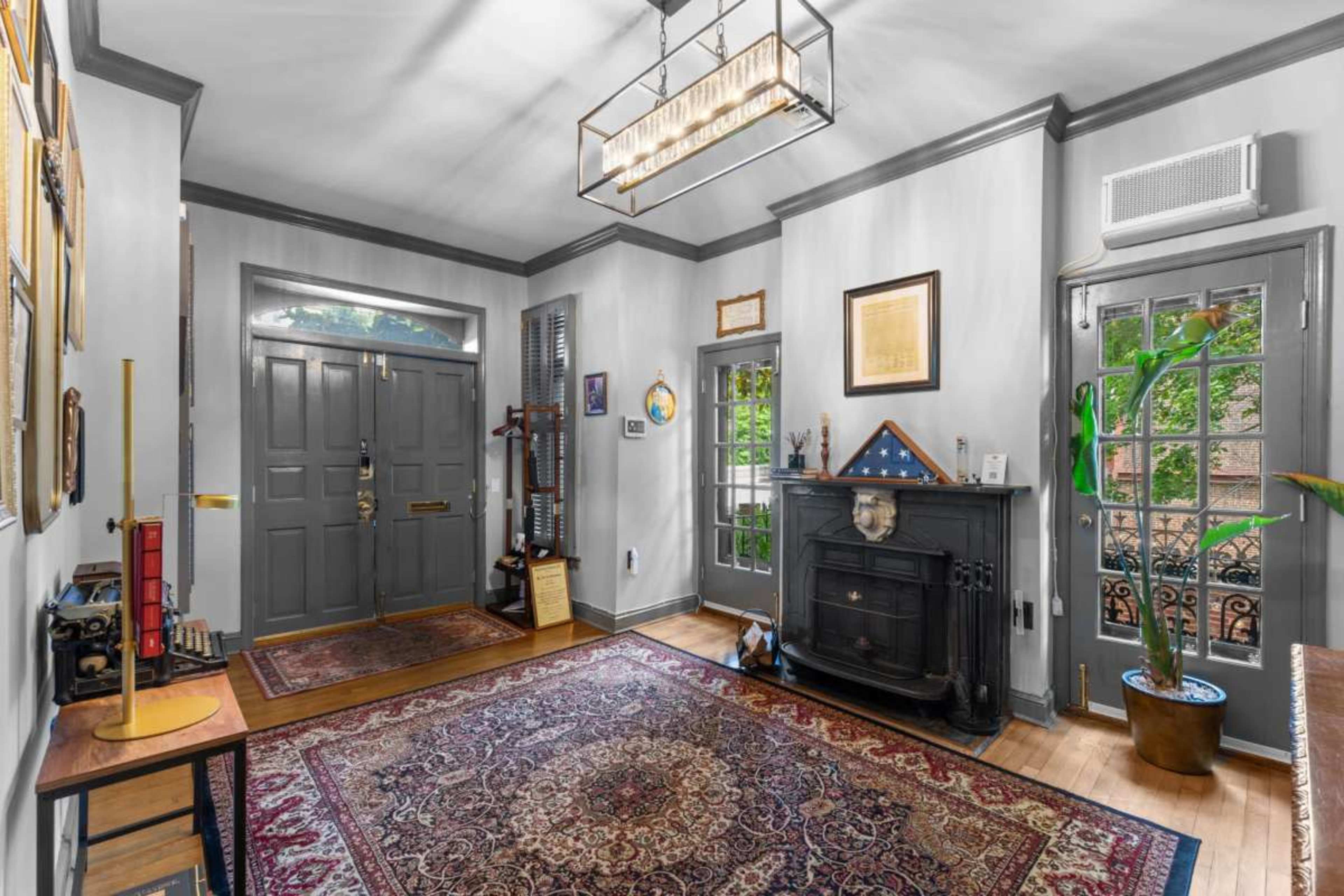 The image shows a well-lit hallway featuring a decorative rug, a fireplace, and a console table, with doors leading to an outdoor area and a small plant in the corner.