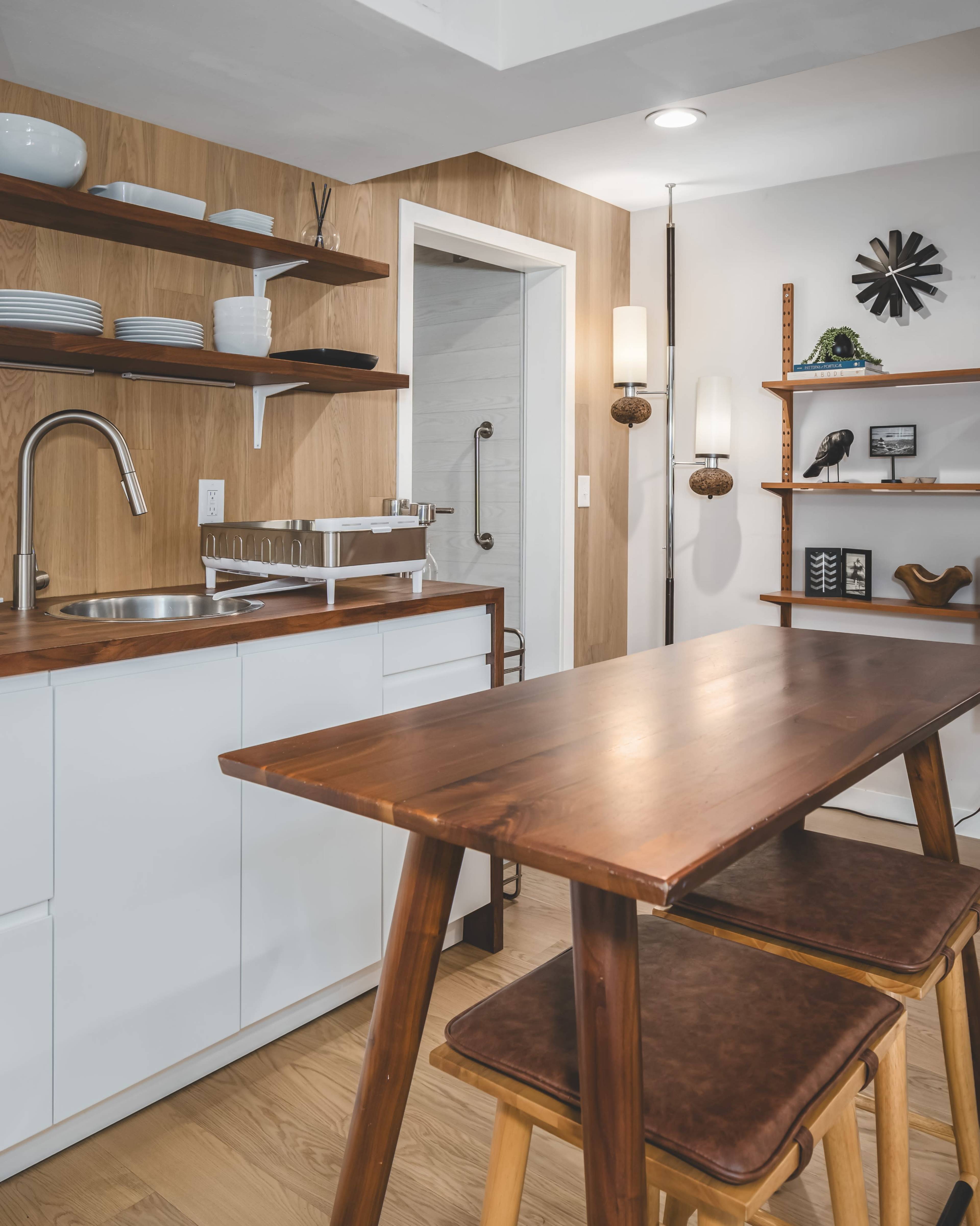 The image features a modern kitchen with wooden shelves, a sleek sink, and a dining table with two stools.