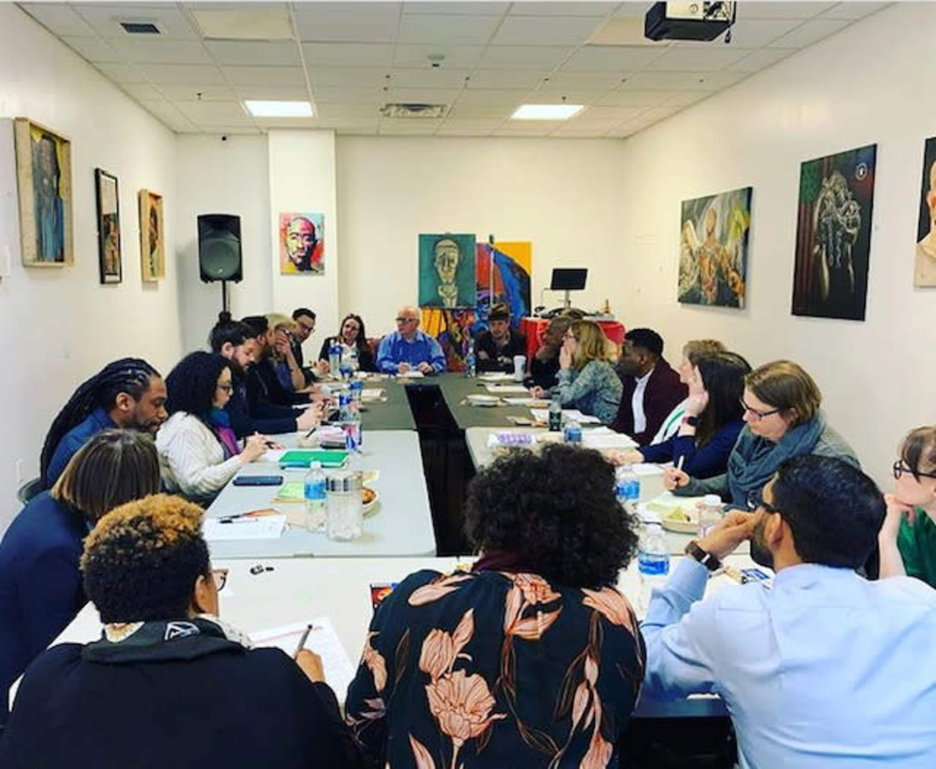 A diverse group of people is seated around a long table in a well-lit conference room, engaged in discussion while various artworks adorn the walls.