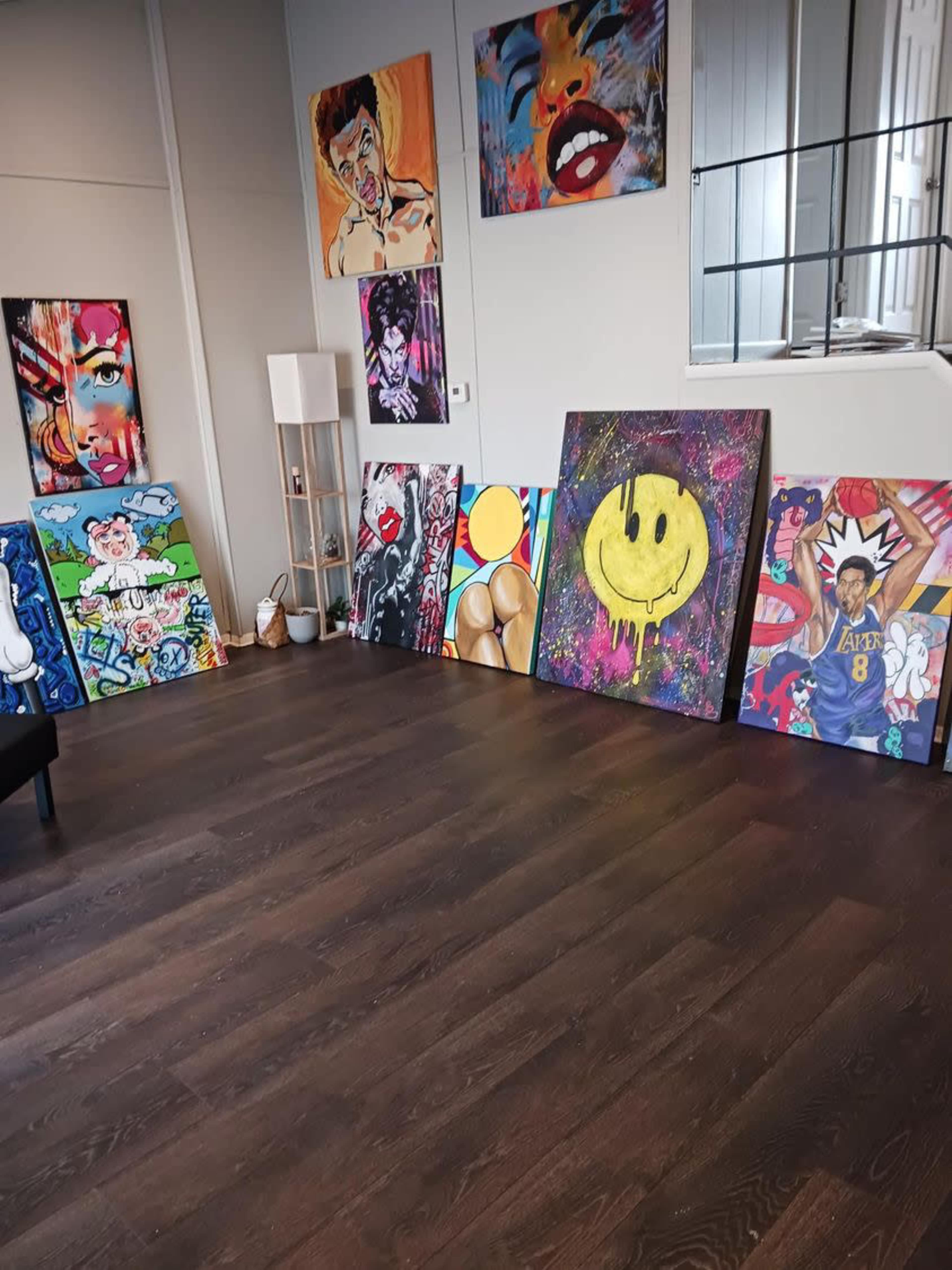 An art studio with several colorful paintings displayed on the walls and propped against the floor, along with a modern lamp and a black chair.