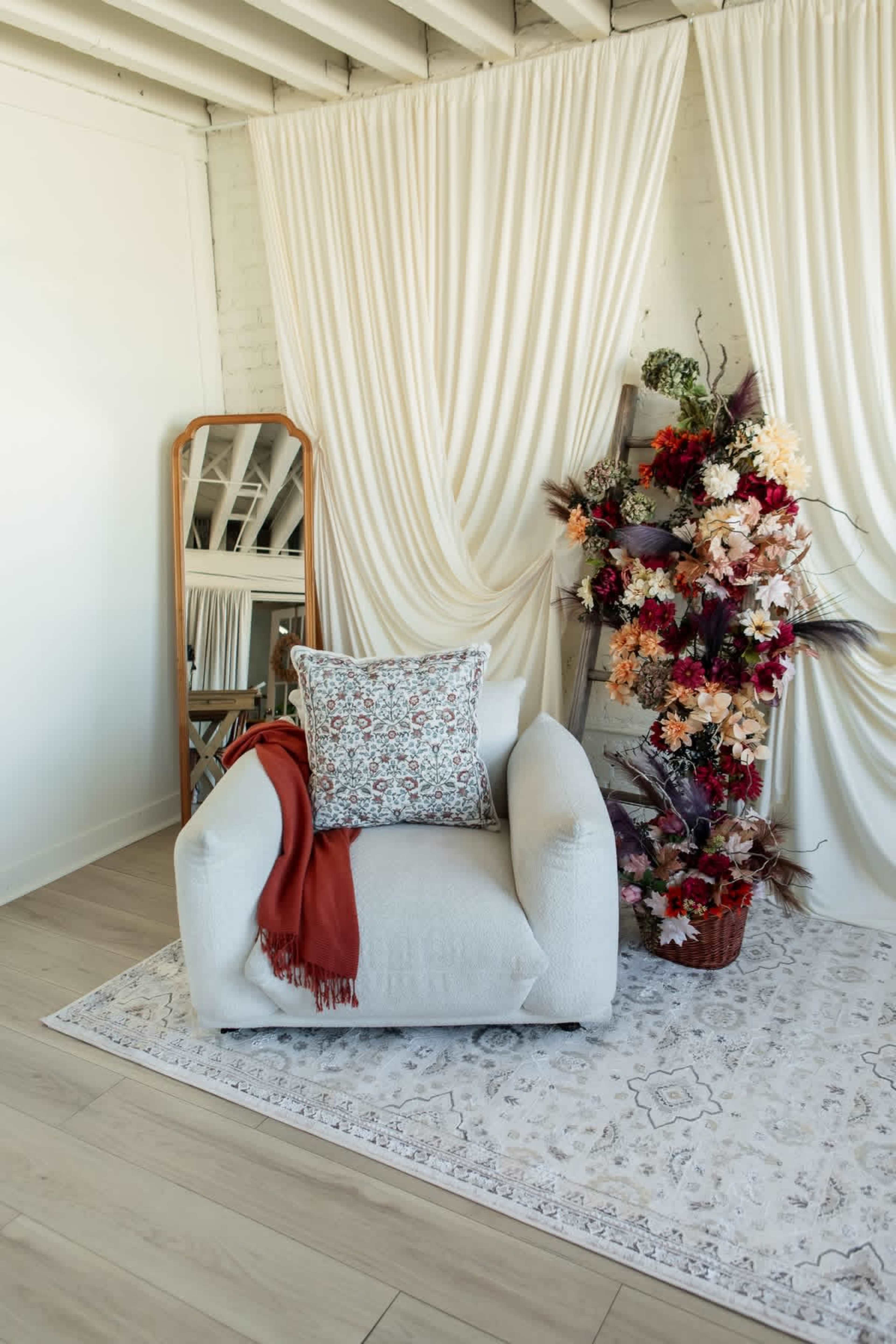 The image features a cozy armchair with a decorative pillow and a red throw blanket, set against a backdrop of draped curtains and a floral arrangement in a decorative pot.