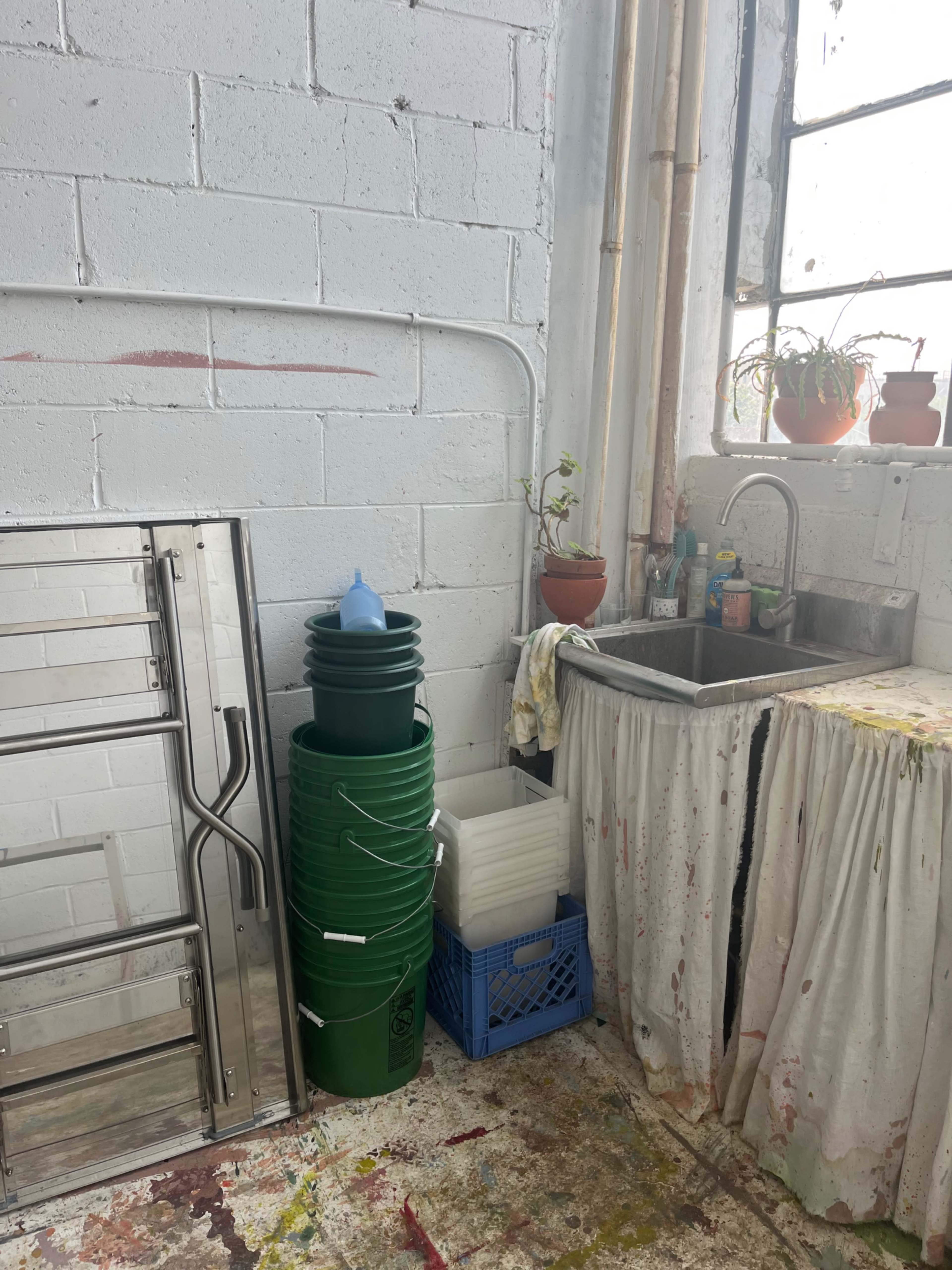 The image shows a small industrial space with a sink, several green buckets stacked beside it, and potted plants on a windowsill.