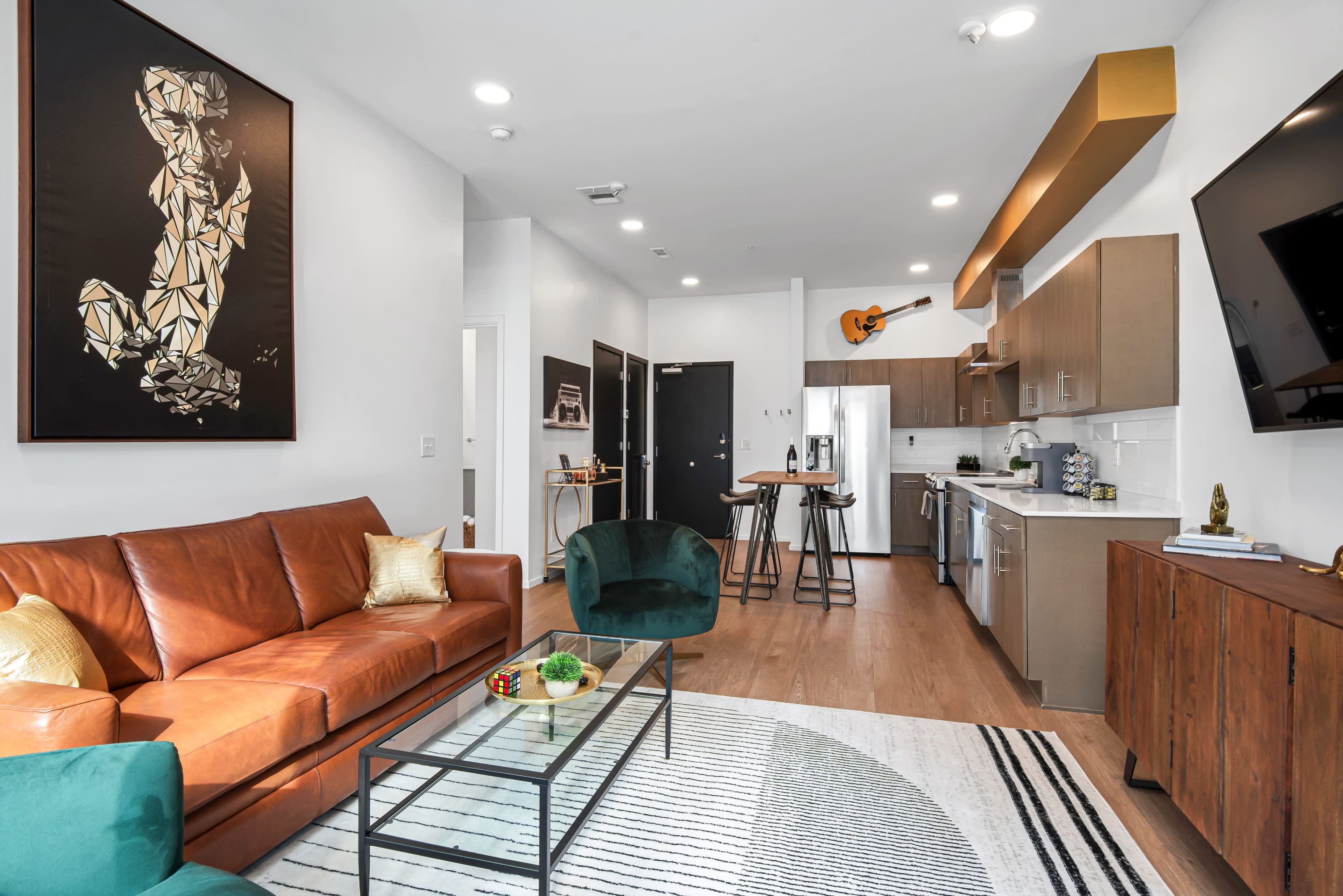 A modern living room with a leather sofa, a glass coffee table, a kitchen area, and a large wall art piece featuring a figure.