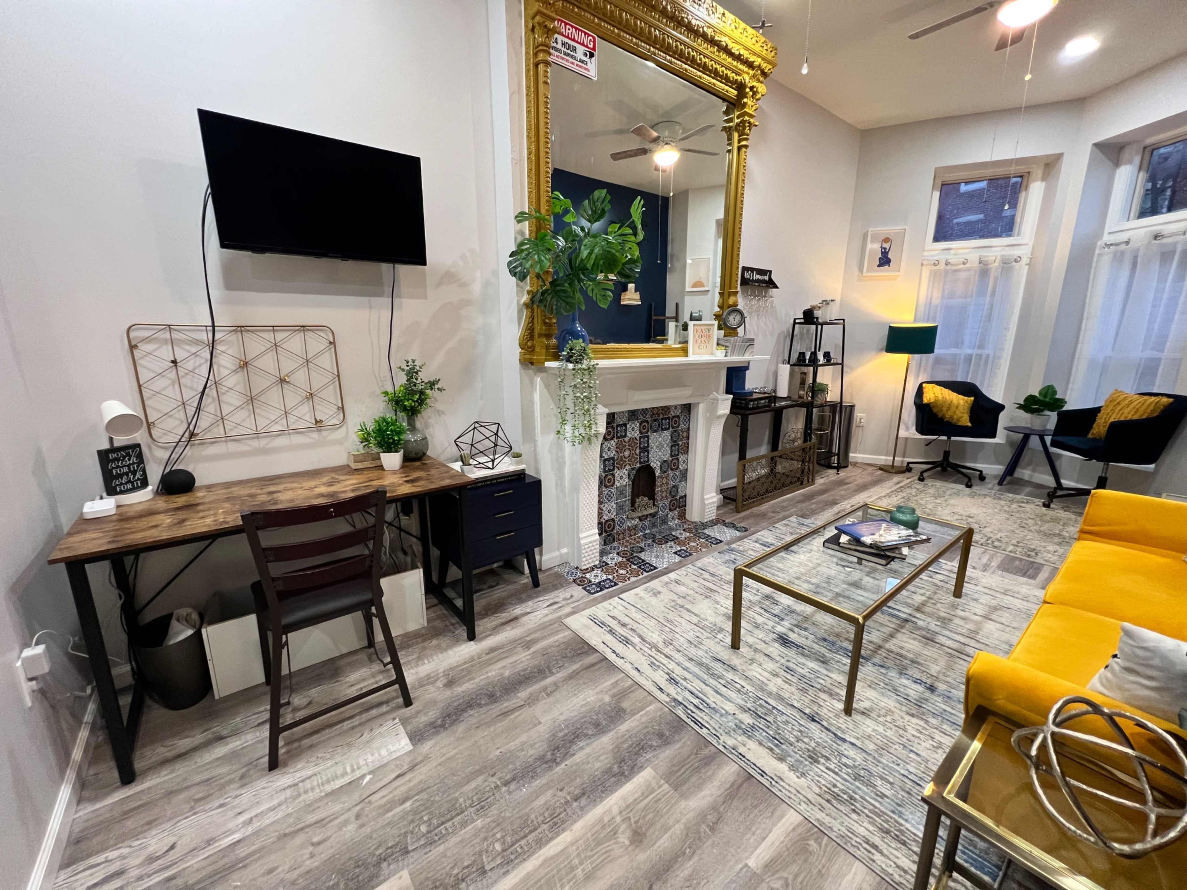 The image shows a stylish living room featuring a yellow sofa, a wooden desk, a decorative fireplace, and large windows with natural light.