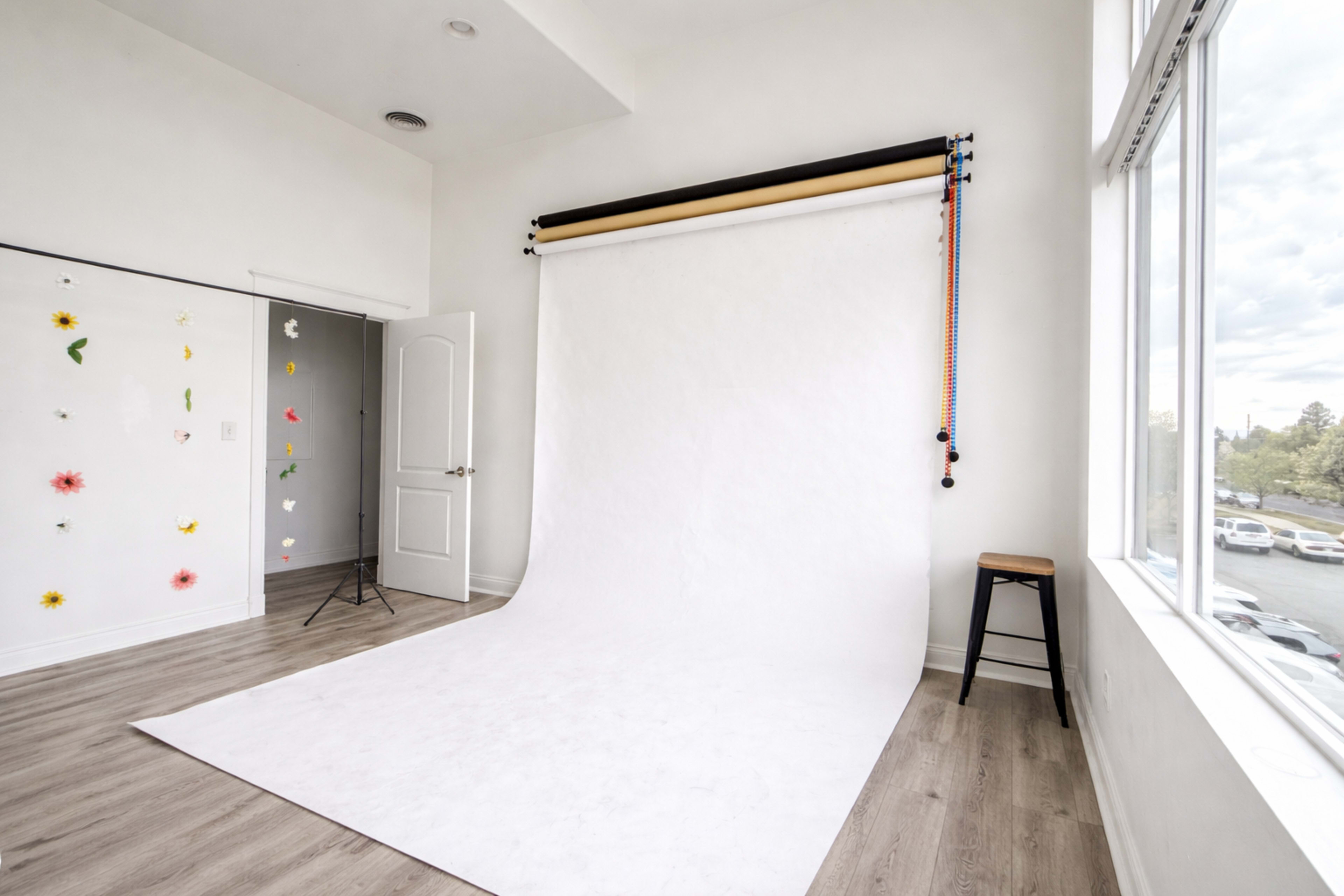 The image shows a photography studio with a white backdrop, light wooden flooring, and colorful decorations on the wall.