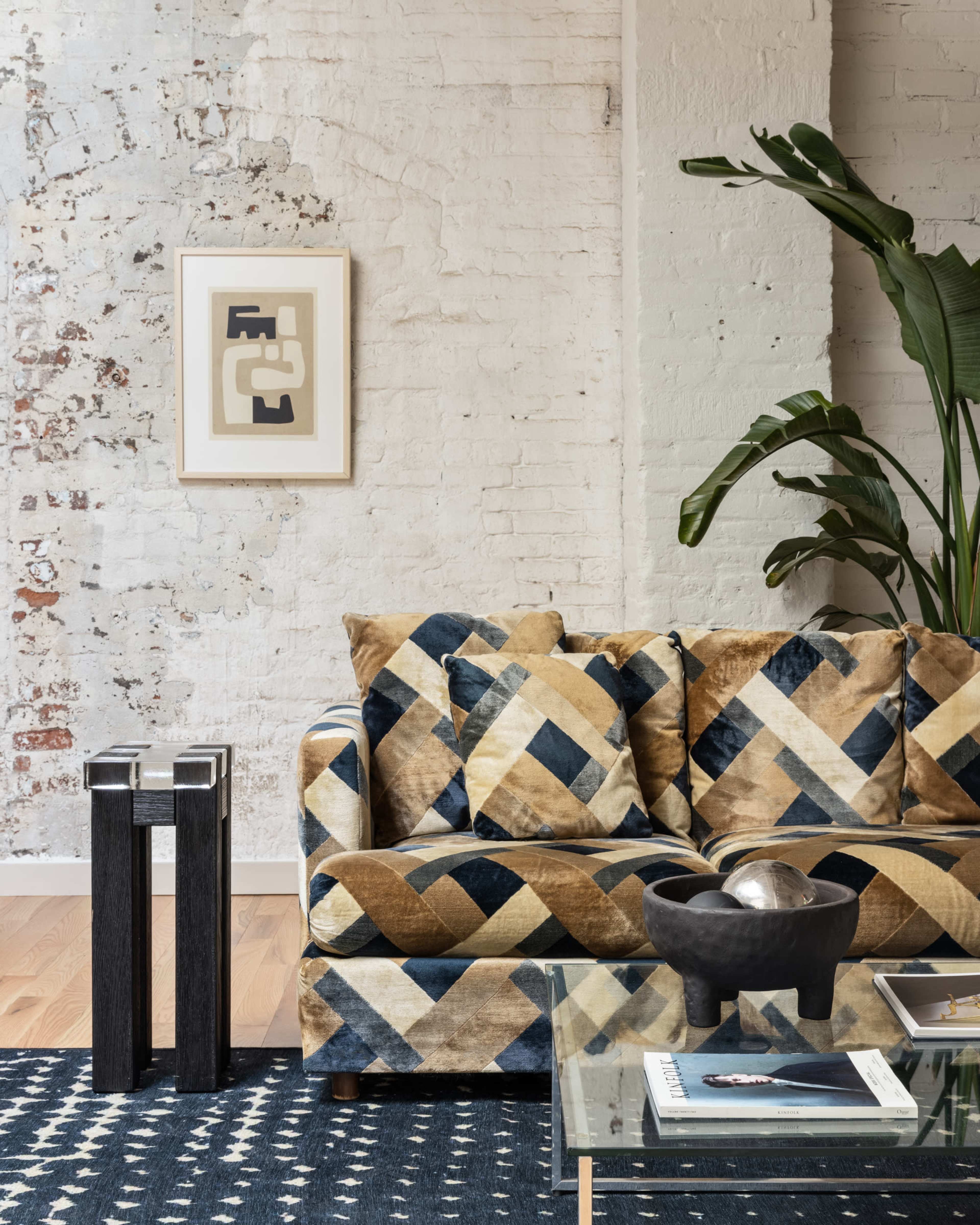 A living room corner with a patterned sofa, a glass coffee table, a black decorative bowl, and potted greenery against a textured white brick wall.