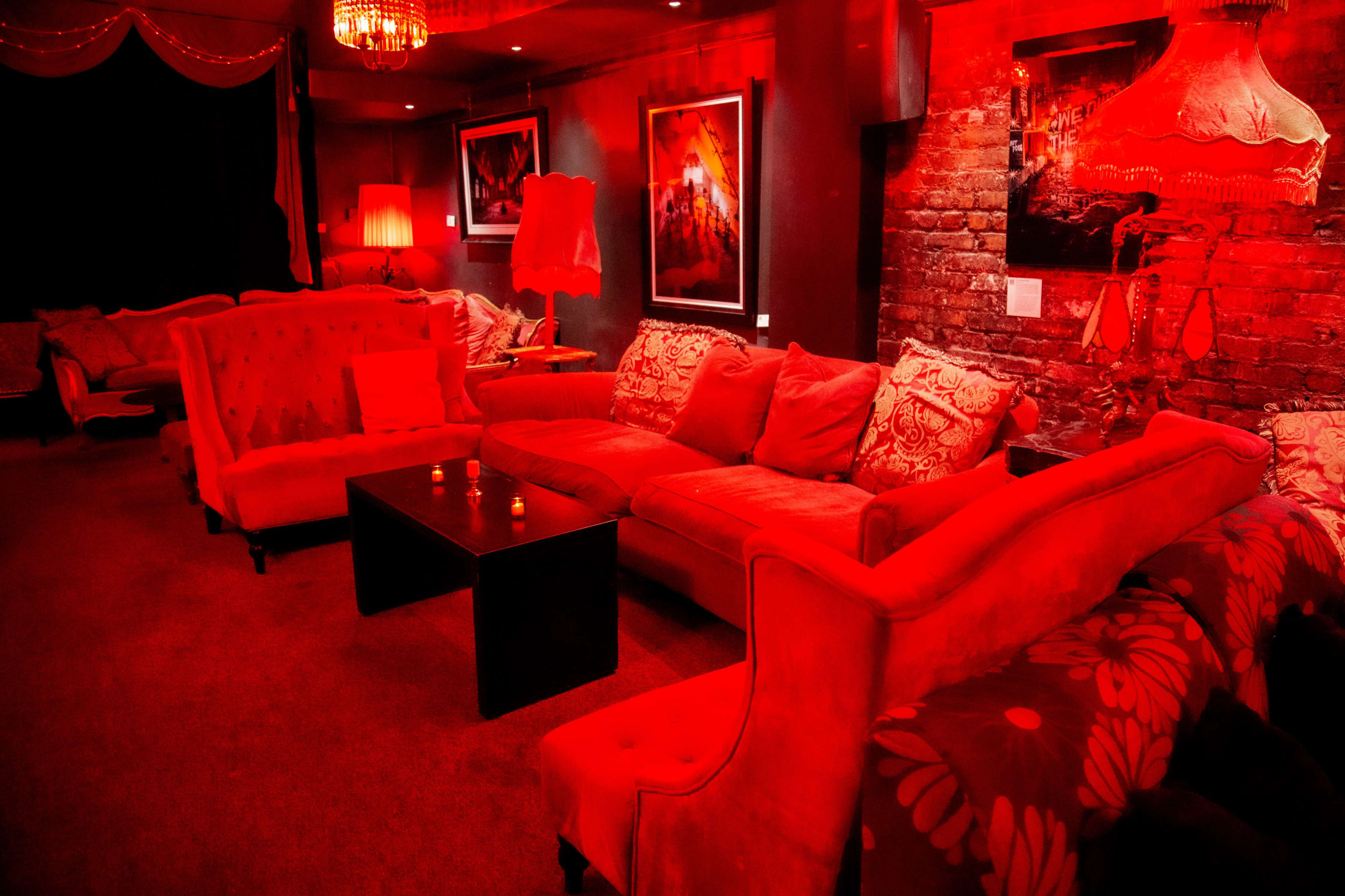 A dimly lit lounge features red sofas, a coffee table, and artwork on the walls against a brick backdrop.