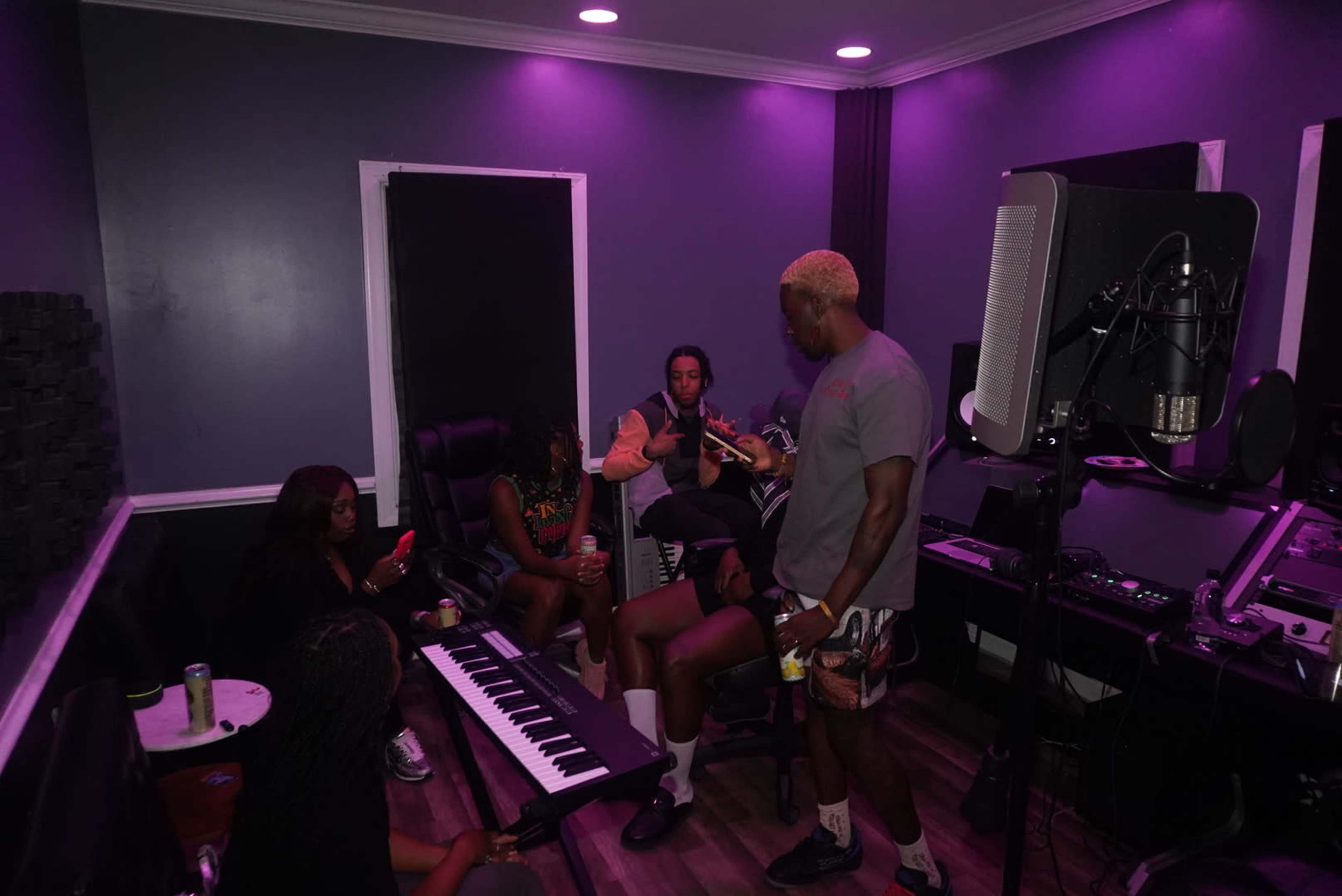 A group of five people is gathered in a music studio with purple lighting, engaged in conversation while a keyboard and recording equipment are present.